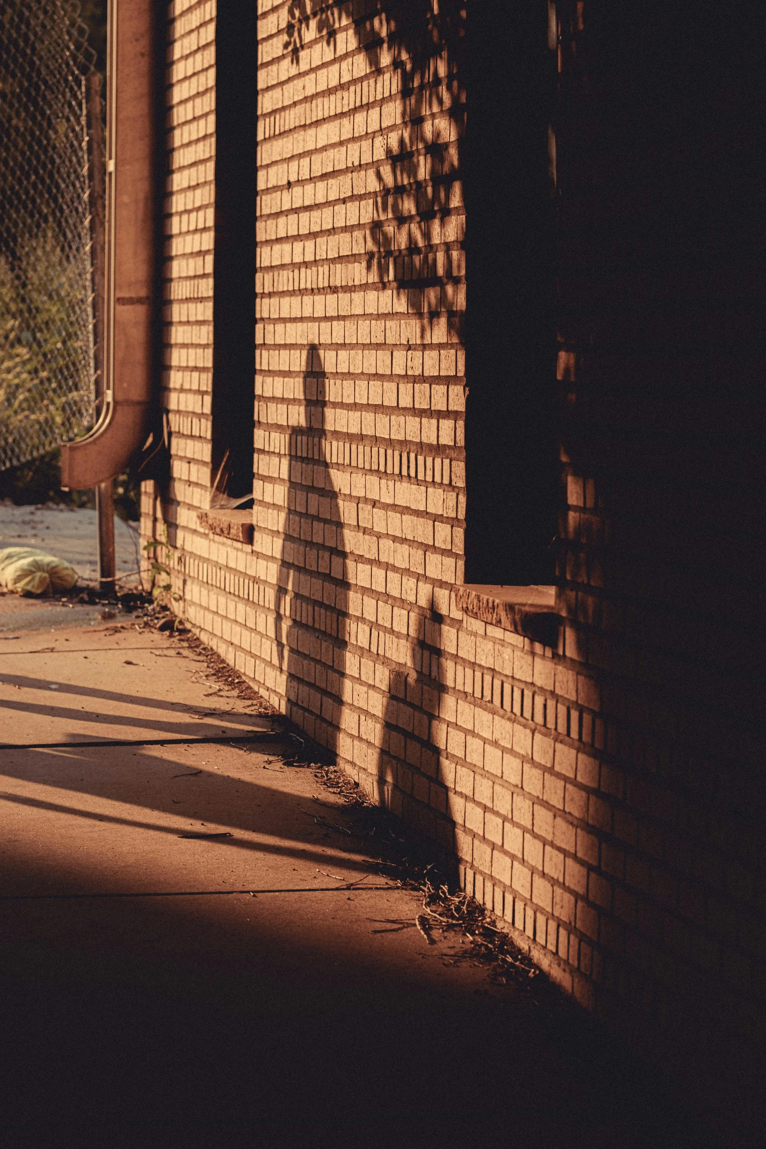Sunset or sunrise casting shadows on a brick wall and sidewalk, with a chain-link fence on the left side.