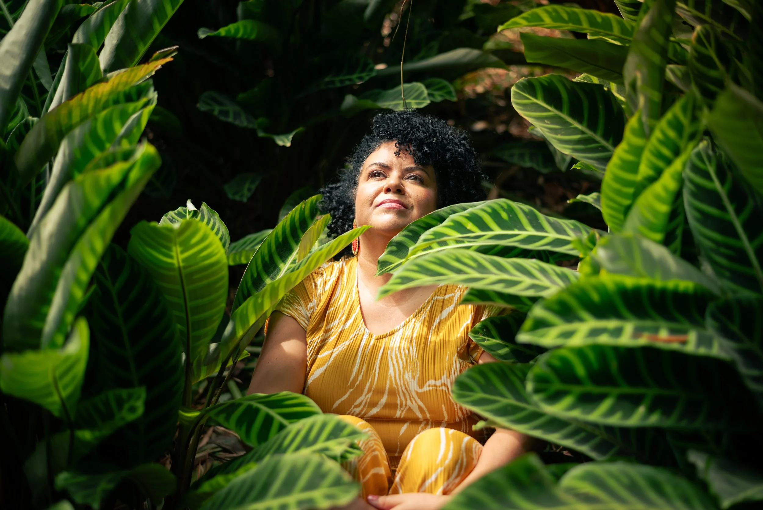 A woman with curly black hair and earrings sitting among green tropical leaves, wearing a yellow patterned dress and looking up.