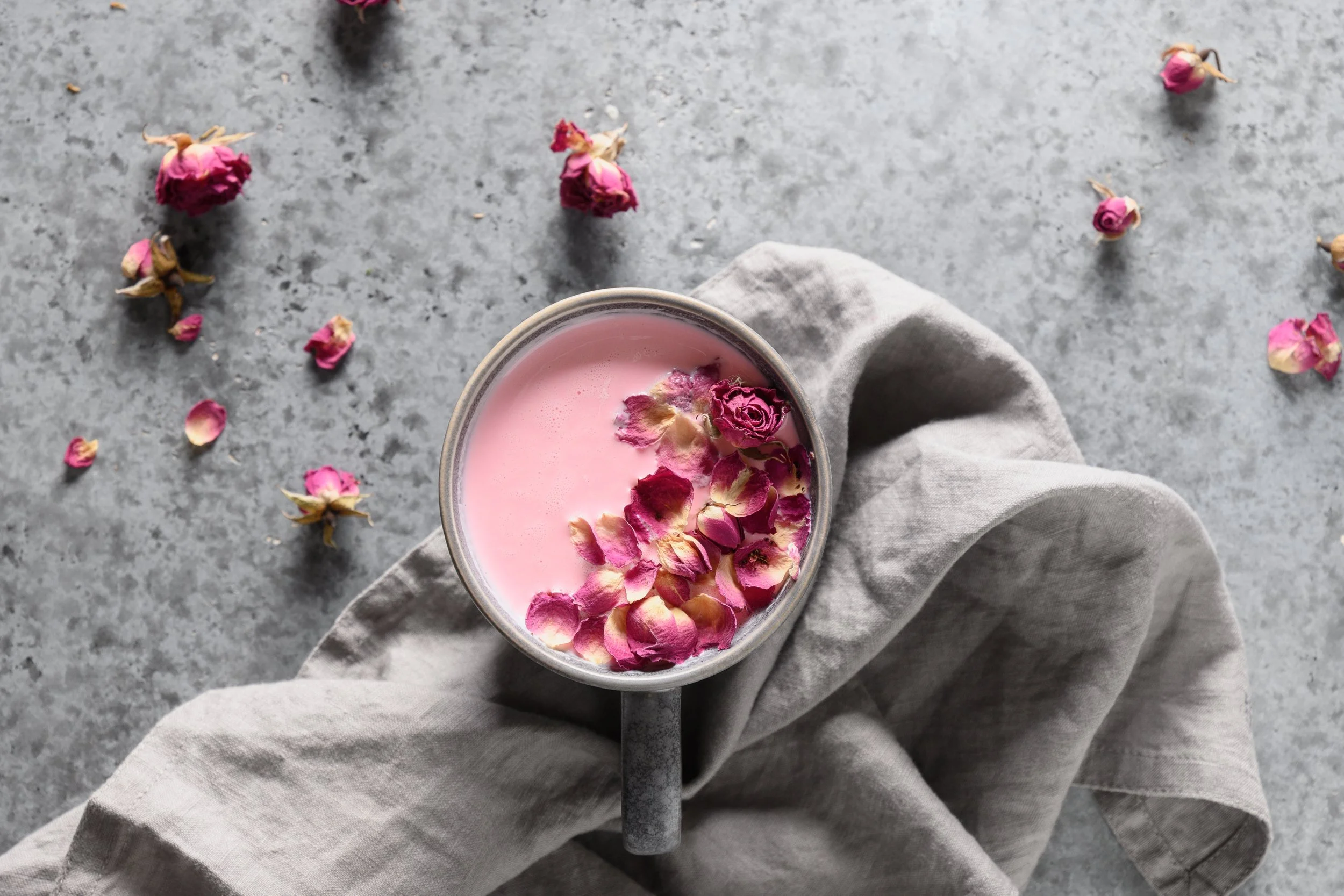 A cup of pink beverage garnished with pink rose petals, placed on a gray textured surface with scattered rose petals around.