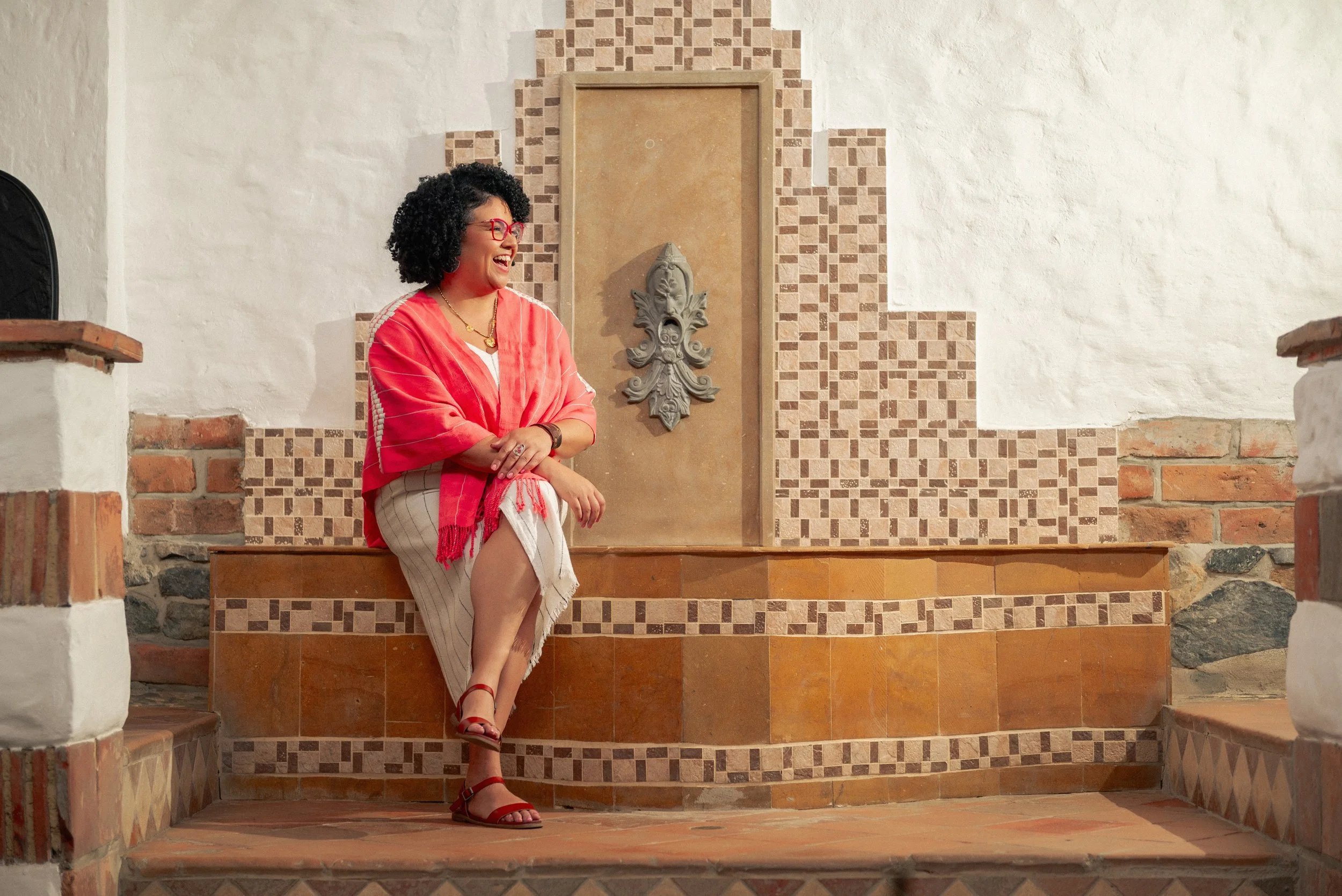Woman with curly black hair and red glasses sitting on steps in front of a decorative wall with a stone face sculpture, dressed in a red shawl, beige skirt, and red sandals, smiling and crossing her arms.