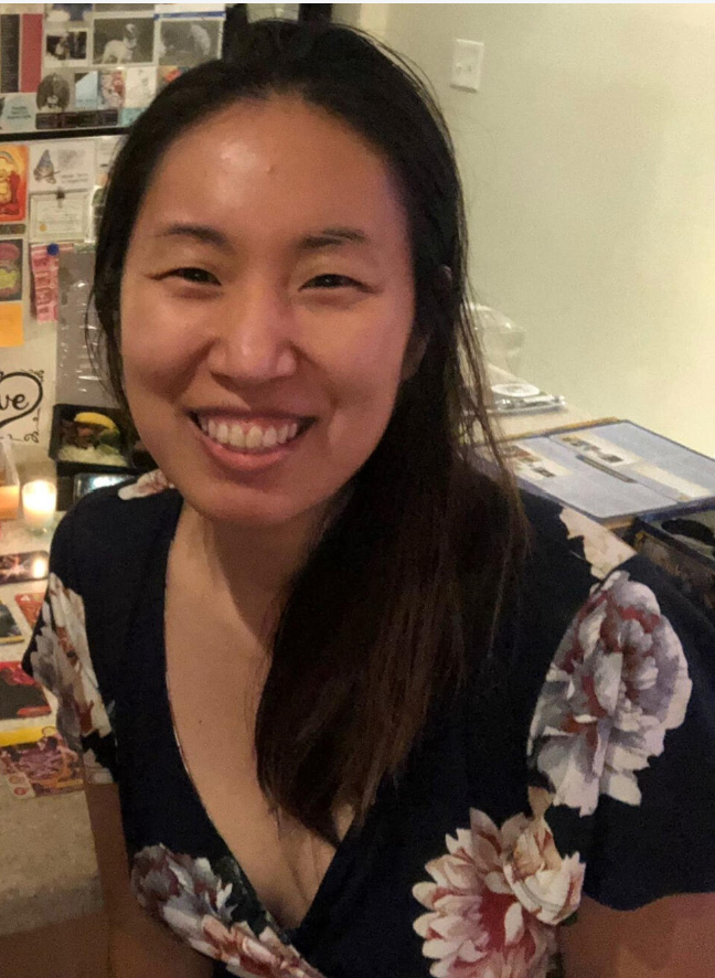 A woman smiling with long dark hair, wearing a black floral dress, in a cozy indoor setting with a wall decorated with photos and postcards behind her.