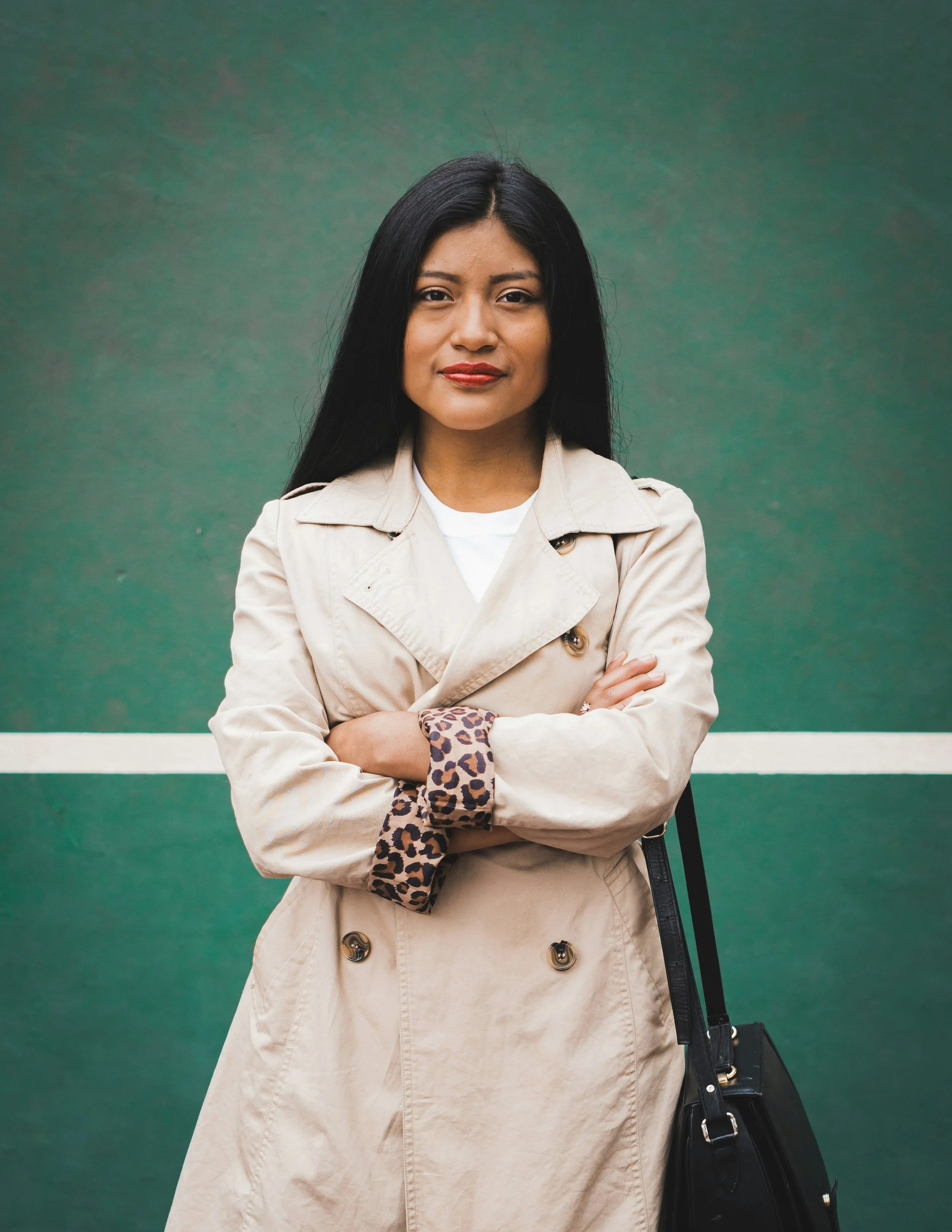 A woman with long black hair, wearing a beige trench coat and a leopard print scrunchie, stands with arms crossed in front of a green background.