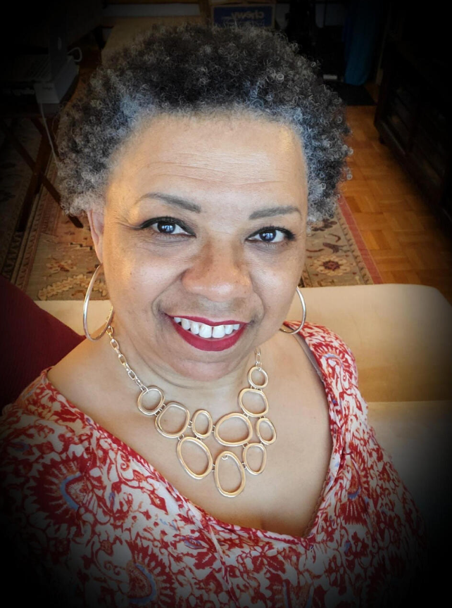 A woman with short gray curly hair, wearing a red and white patterned top, large hoop earrings, and a gold necklace, smiling at the camera in a cozy room with a patterned rug and furniture.