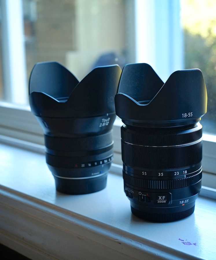 The Zeiss Touit 12mm f/2.8 (Fuji X Mount) in the background compared to the Fuji XF18-55mm zoom in the foreground. About the same length, but features a Zeiss style flare to a significantly wider front element. 