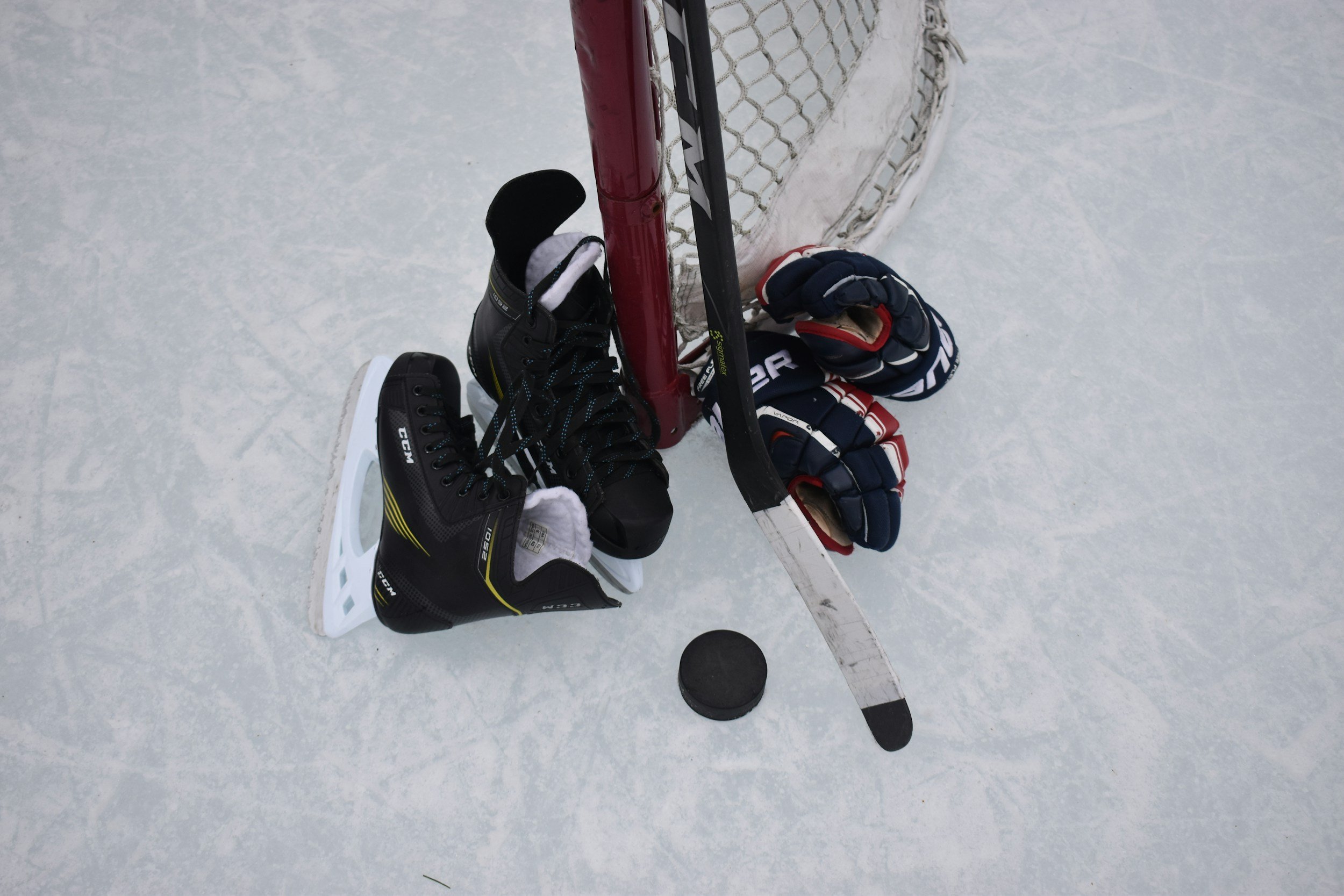 Hockey skates, gloves, a stick, and a puck leaning against a net on ice