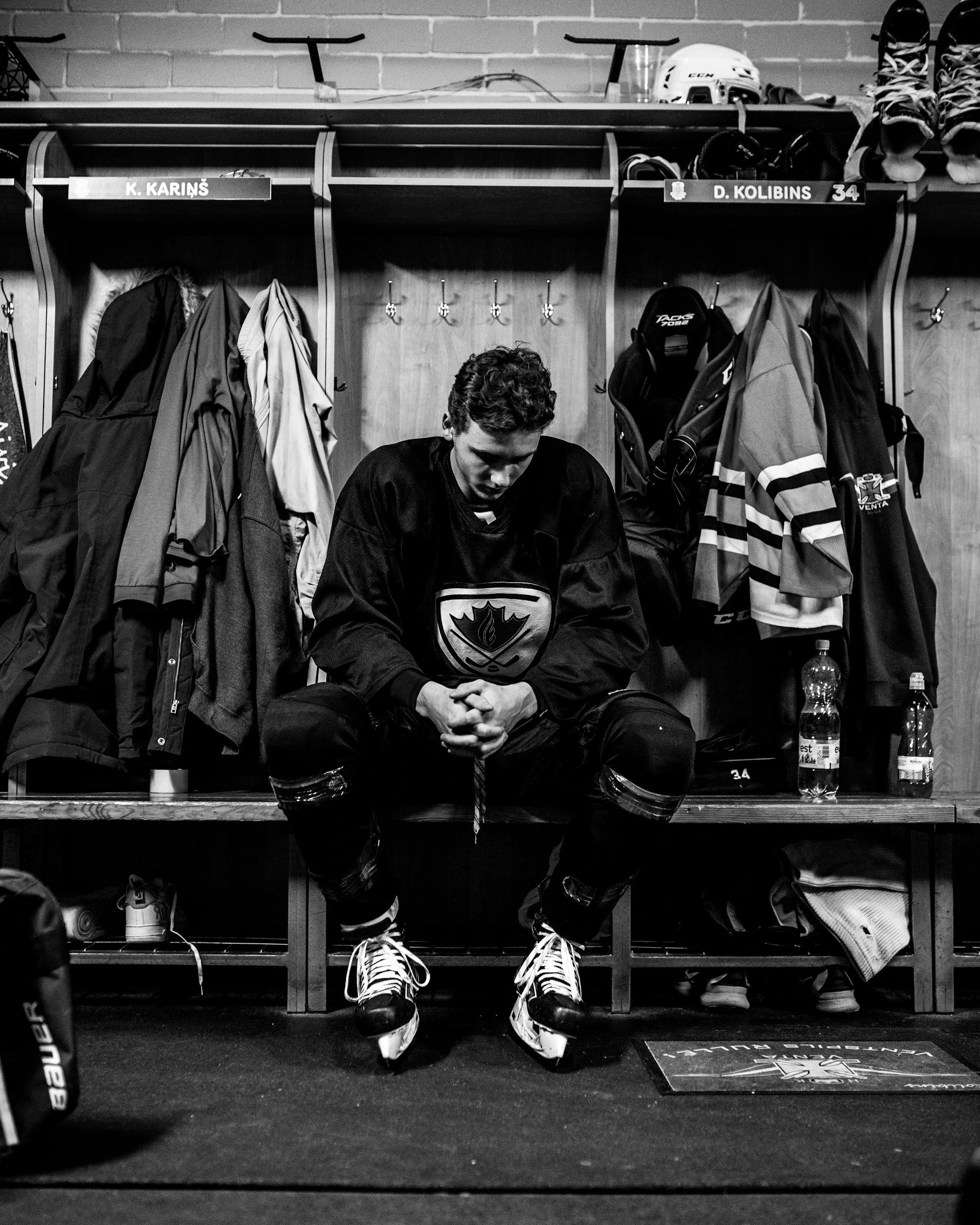 Hockey player sitting in the locker room in his gear with his hands folded, looking down at his feet.