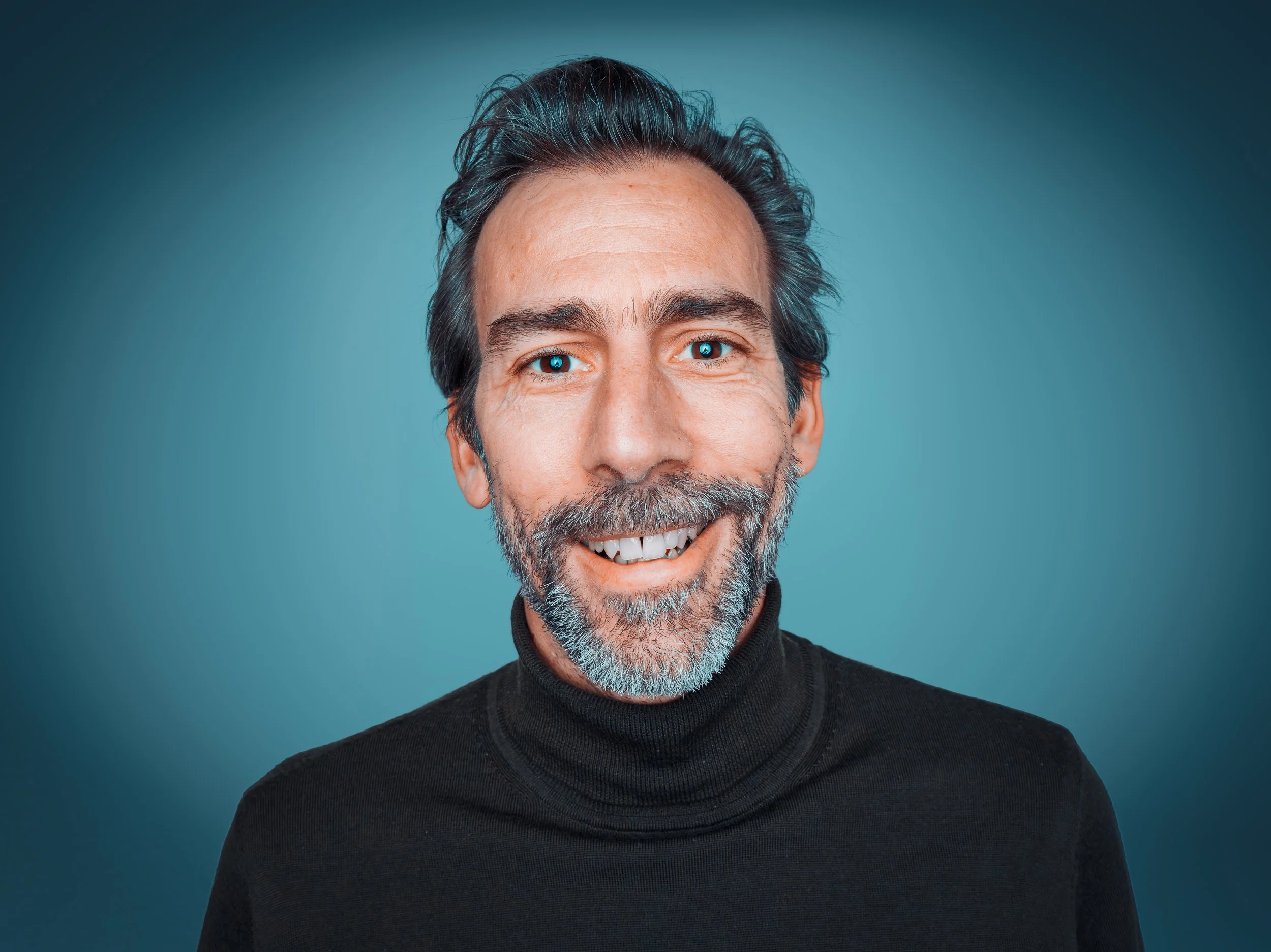 A smiling man with gray hair, glasses, and a blue striped shirt, holding a glass of white wine, with a playful expression and head tilted towards the glass against a dark background.