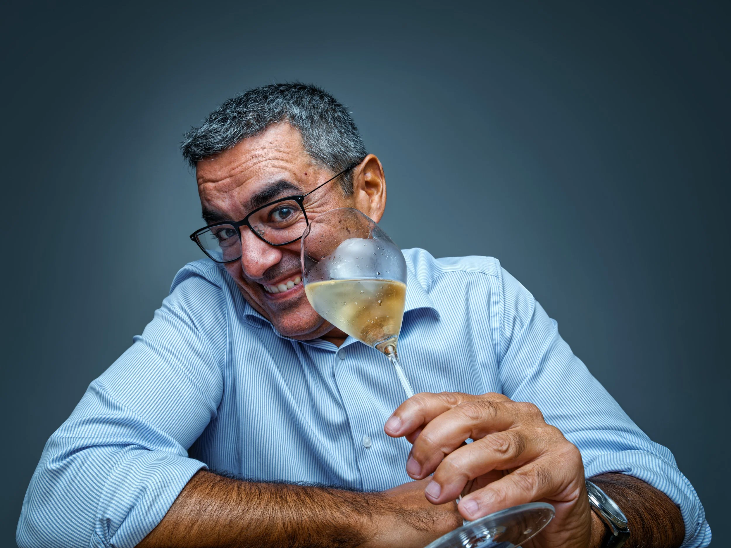 A smiling man with gray hair, glasses, and a blue striped shirt, holding a glass of white wine, with a playful expression and head tilted towards the glass against a dark background.