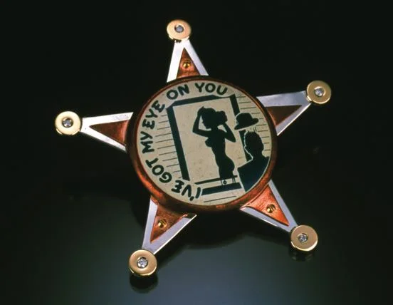This brooch is a humorously critical view of the overreaching Patriot Act. 14 karat gold discs with flush set diamonds are soldered to a sterling silver base, while a tarnished copper bezel secured with brass hardware holds the punchline in the form of an antique pinback.