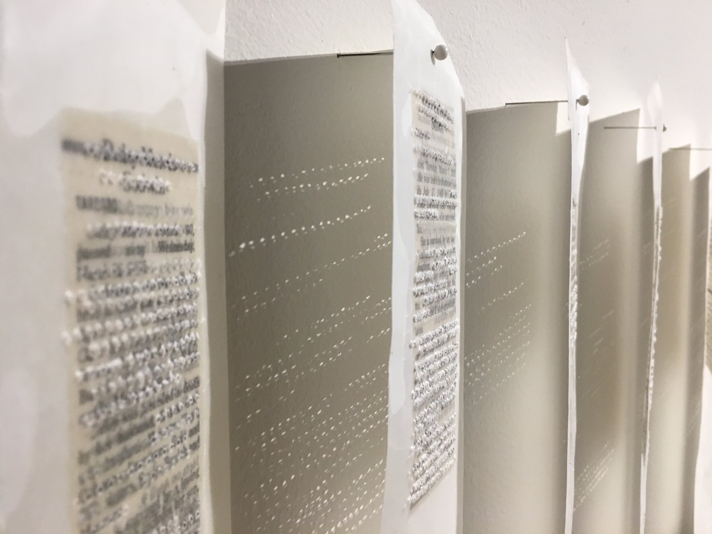 detail of obituaries encased in paper, waxed. holes visible. light filtering through holes onto gallery wall.