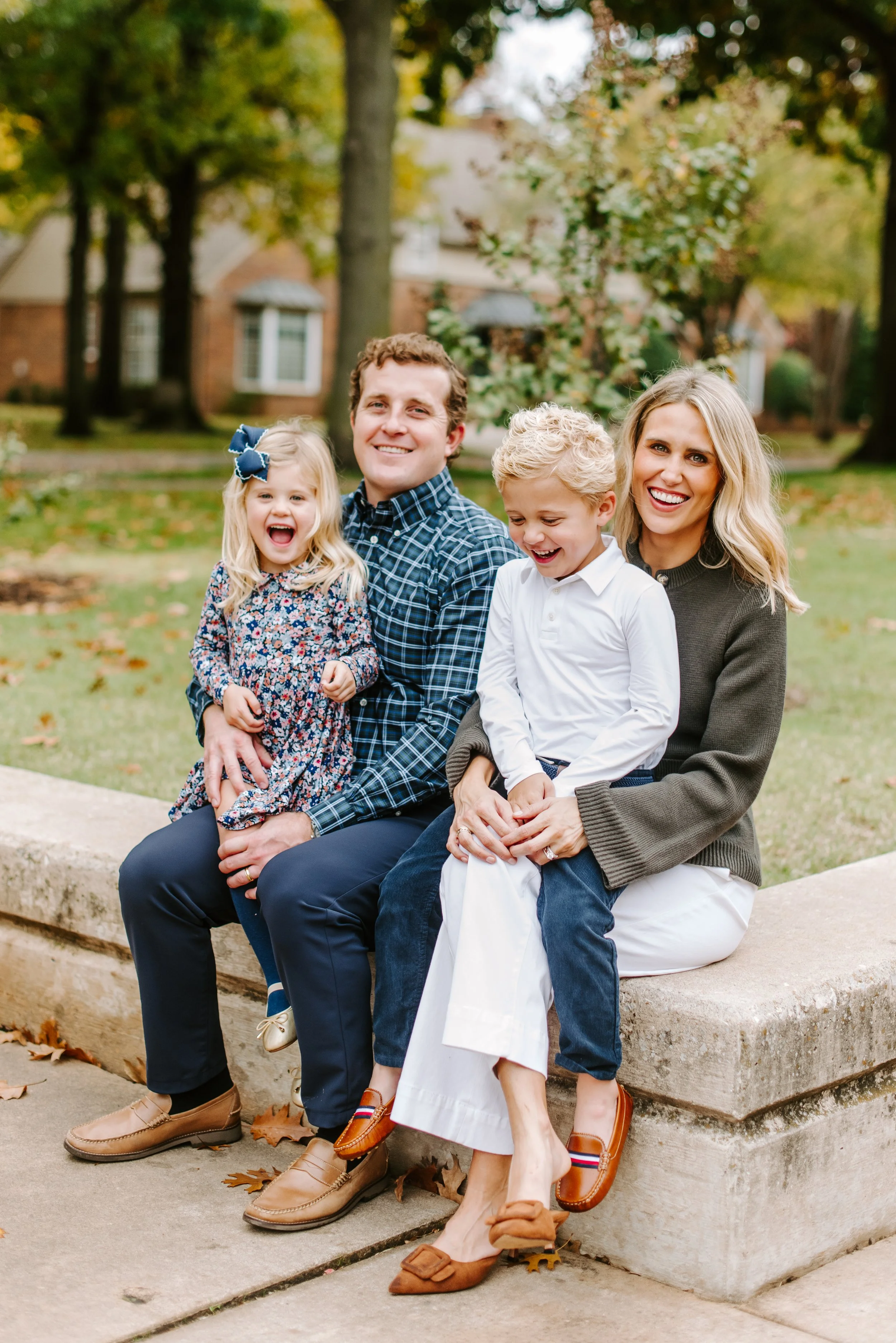 A family of four sitting on a stone ledge outdoors in a park during autumn, smiling and laughing. The family includes a young girl with a bow in her hair, a man, a boy, and a woman. The background shows trees with fall foliage and a house.