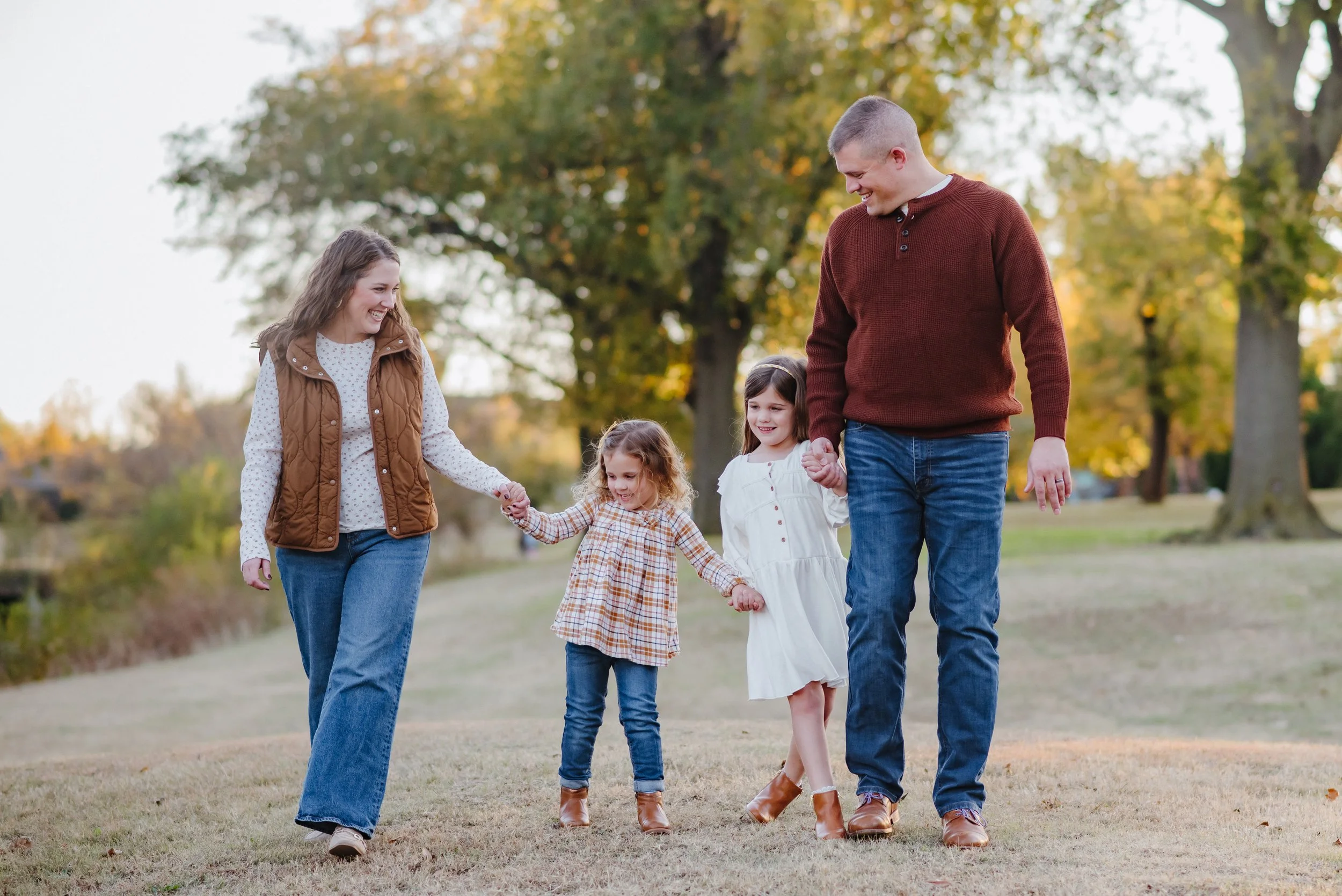 A family of five, including a mother, father, and three young girls, walks hand-in-hand through a park with fall foliage in the background, smiling and enjoying the outdoor autumn day.