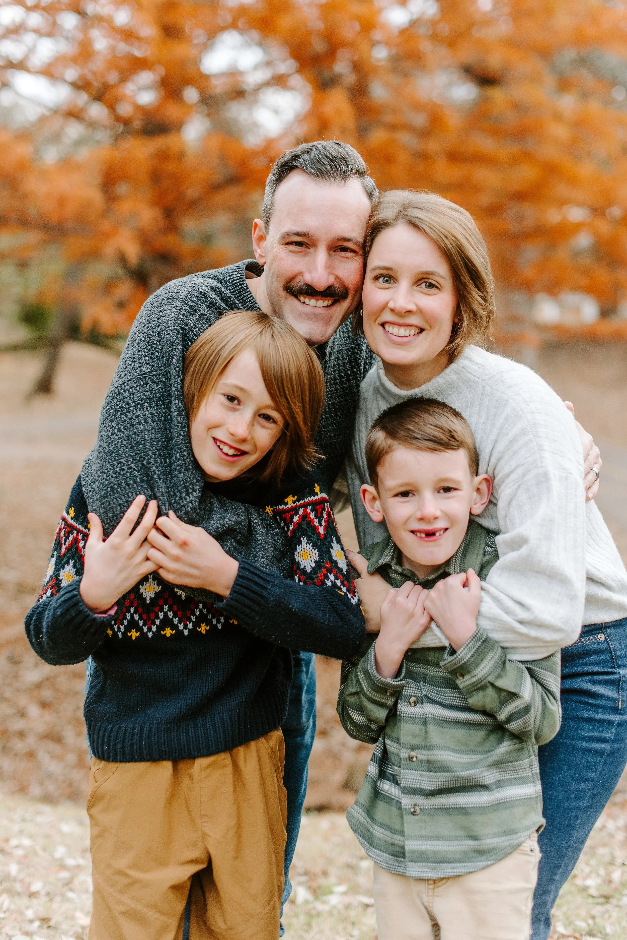 Family of five smiling and hugging outdoors in autumn, with orange leaves in the background.