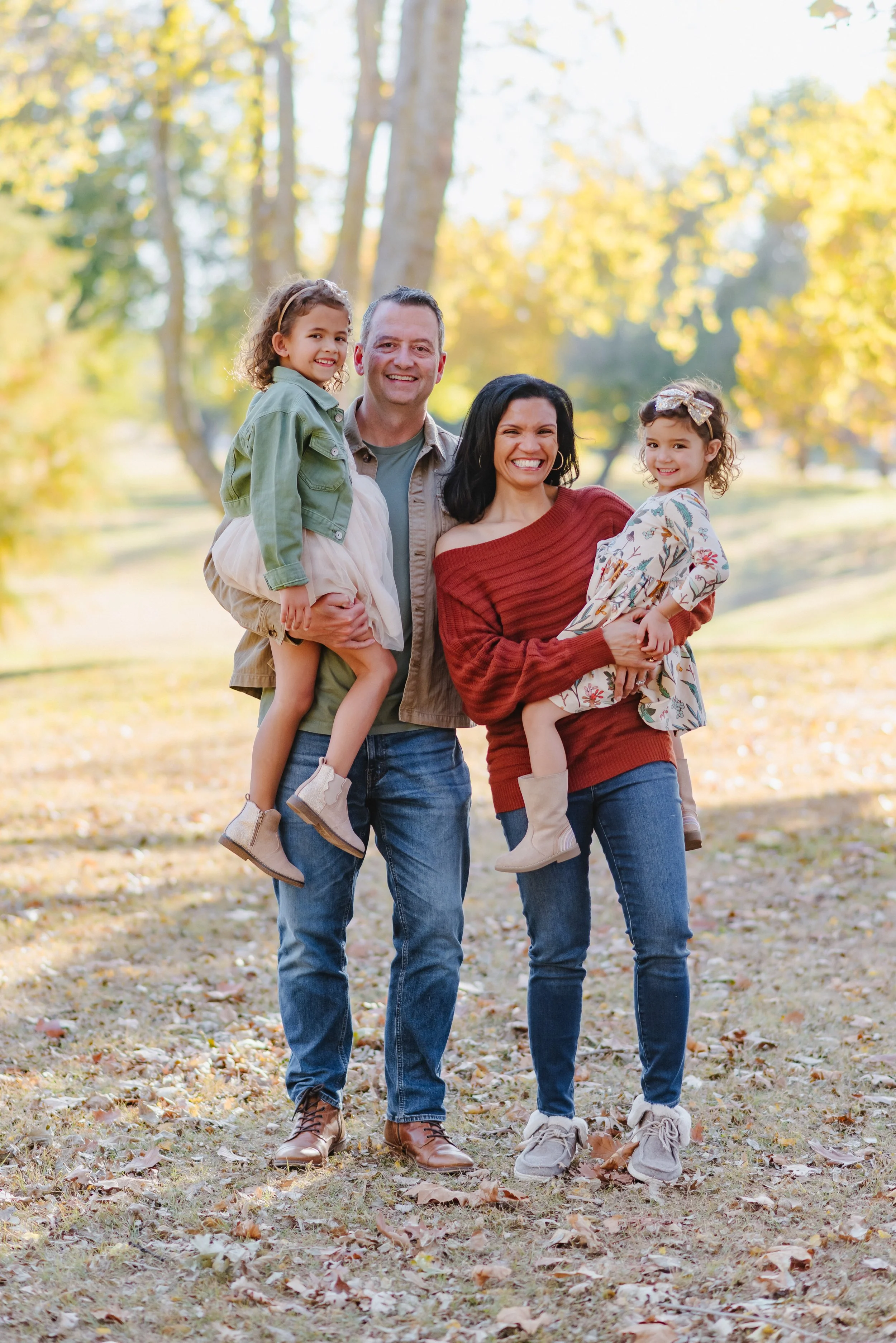 A happy family of four walking outdoors in a park during autumn. The father is holding his daughter and the mother is holding her younger daughter. They are all smiling and wearing casual clothing.