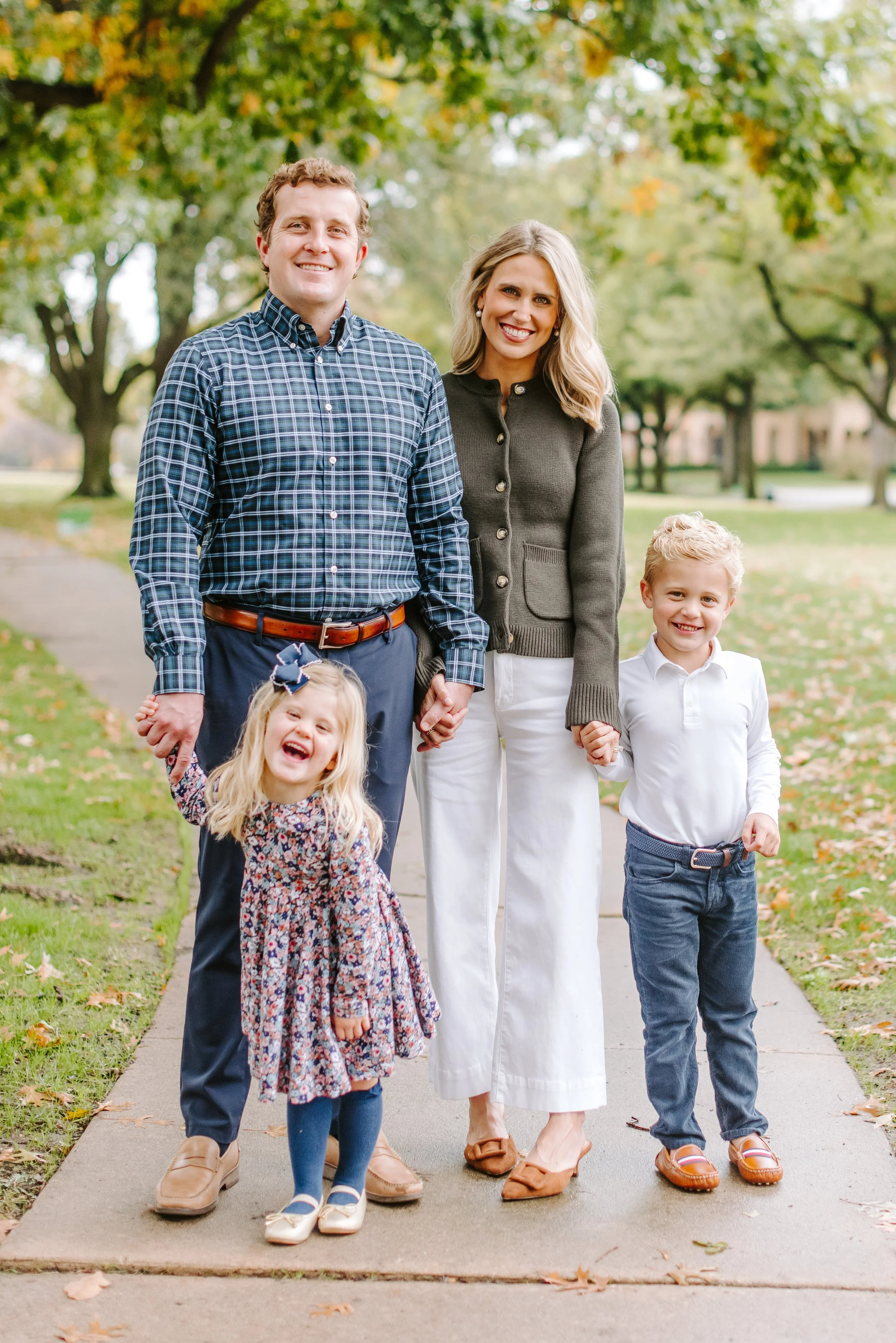 A family of five holding hands and walking on a sidewalk in a park with trees and fall foliage, smiling at the camera.