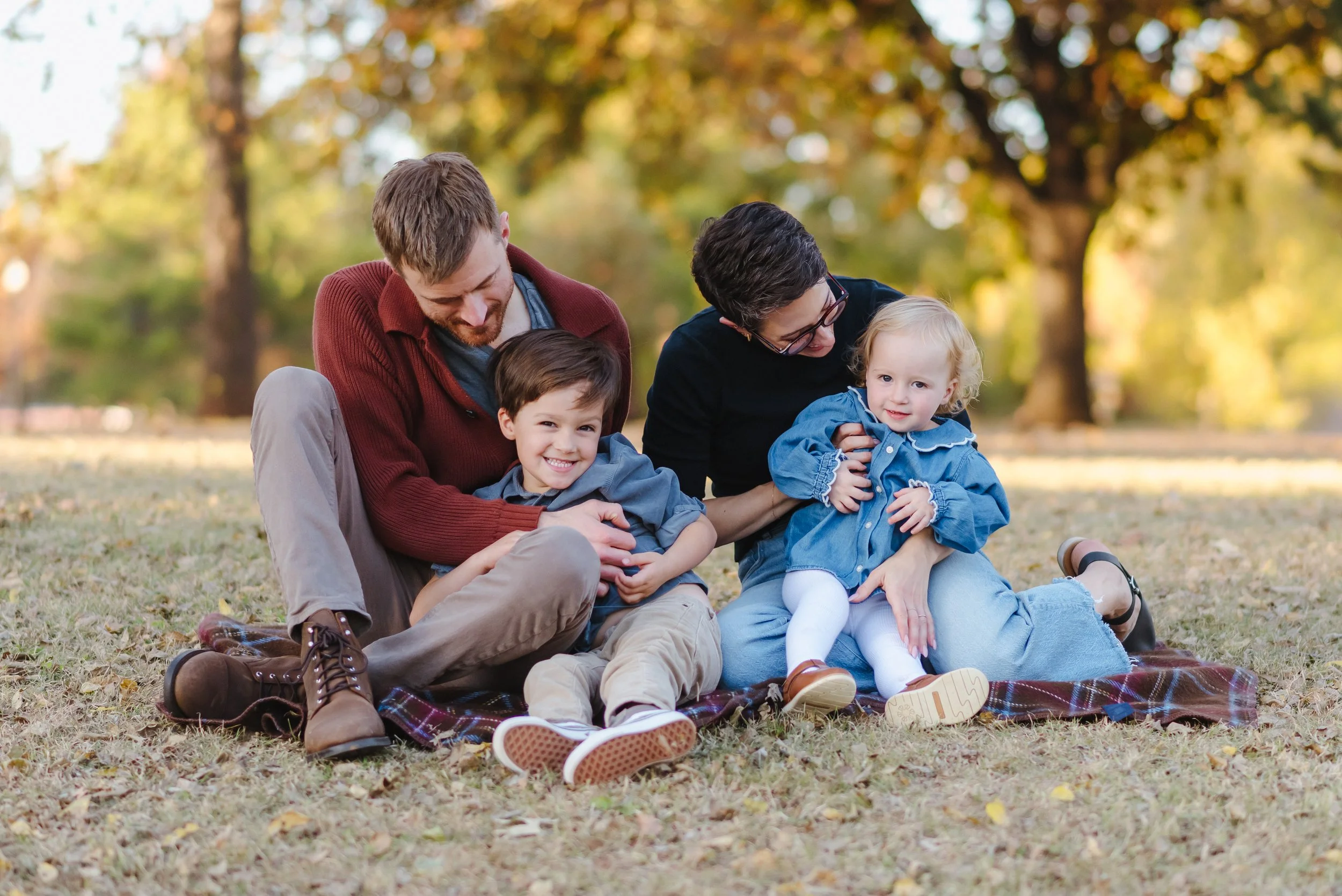 A family of four sitting on a blanket in a park during autumn. The father and mother are holding two young children, with trees and fall leaves in the background.