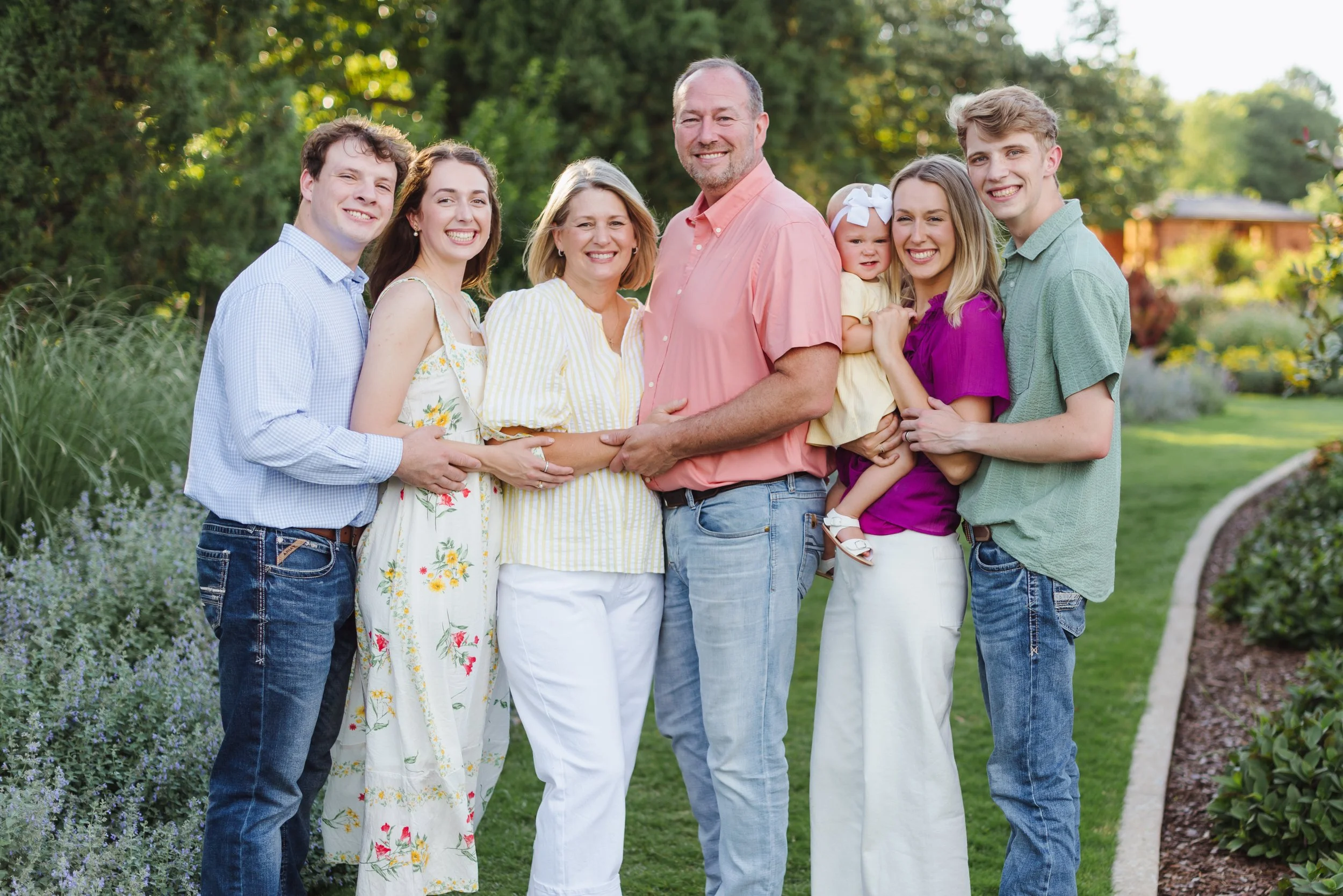 A multigenerational family standing outdoors in a park, smiling at the camera, with trees and greenery in the background.