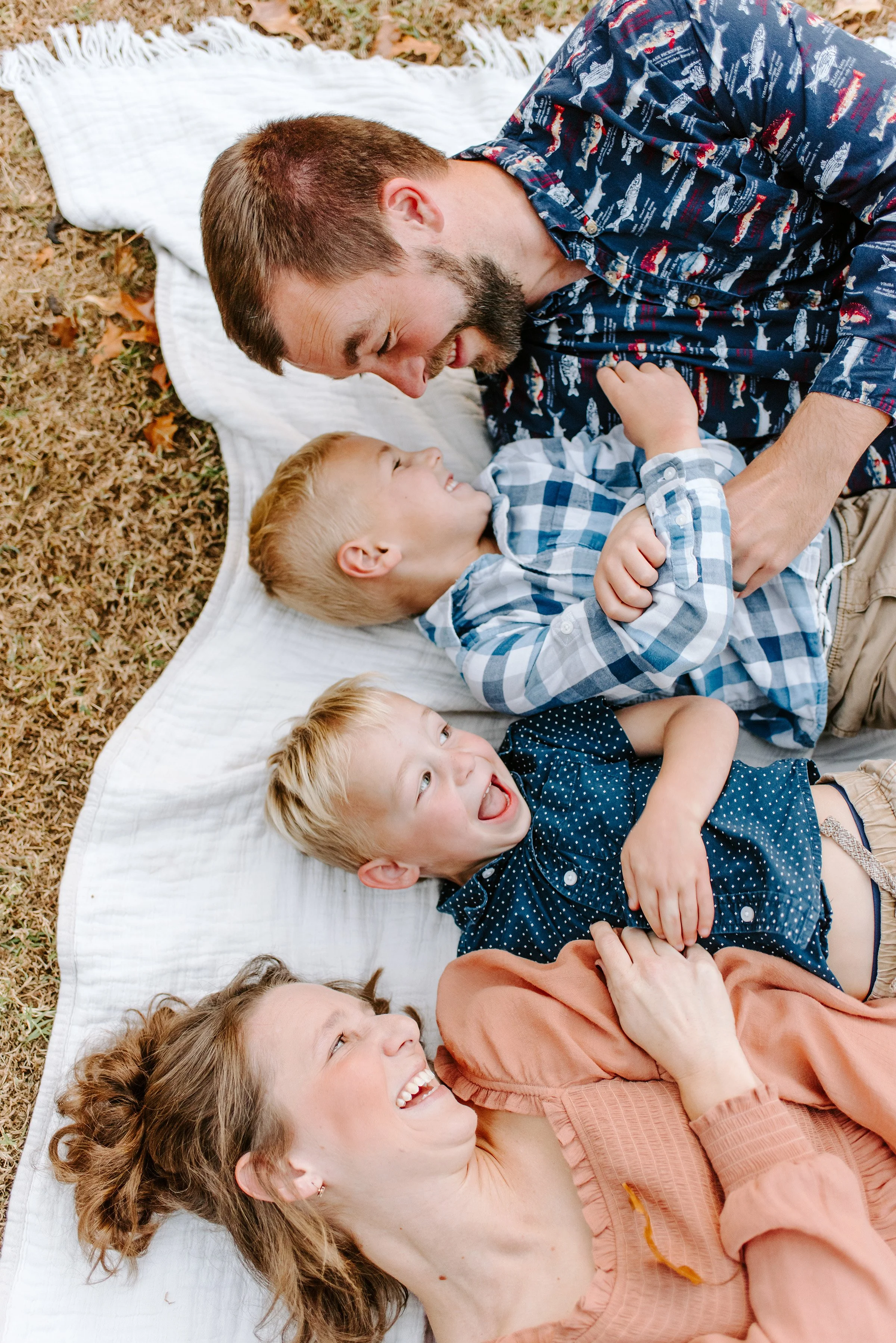 A family lying on a white blanket on the grass outdoors, smiling and engaging with each other.