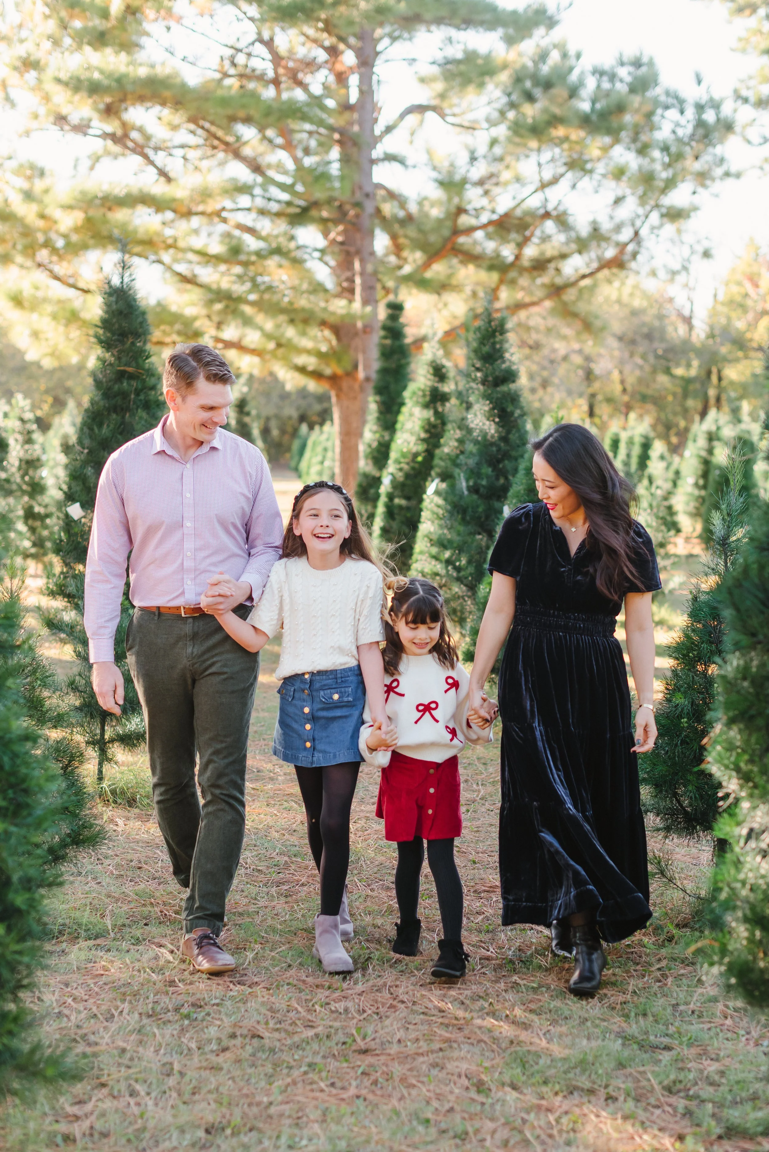 Family walking through a Christmas tree farm, holding hands and smiling, with tall evergreen trees in the background.