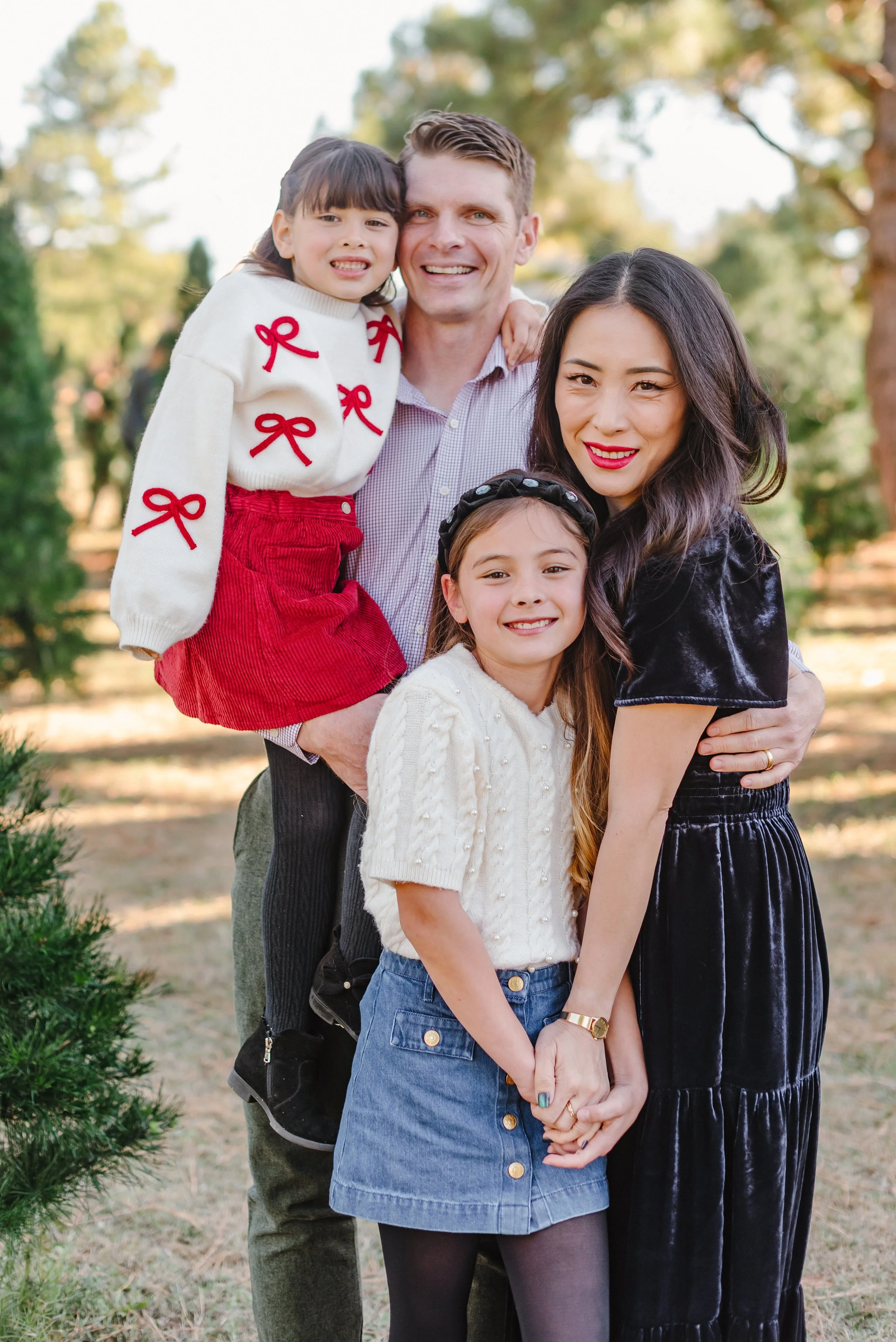 A family of four smiling outdoors with a woman, man, and two young girls, one of which is wearing a sweater with red bows