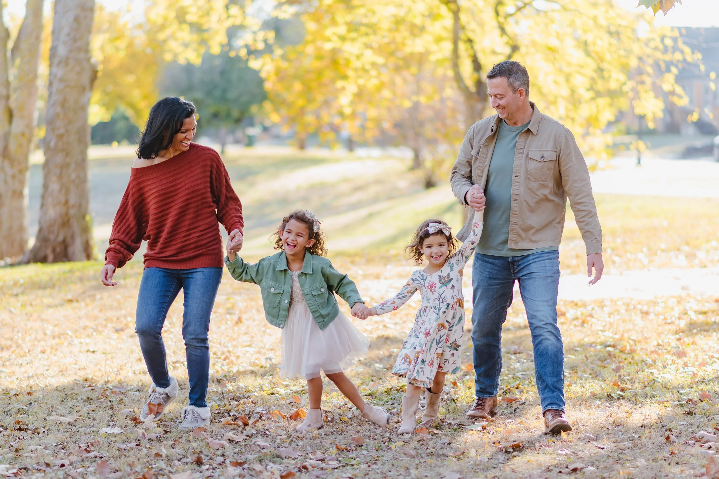 A family of four walking through a park with autumn leaves on the ground, smiling and enjoying a sunny day.
