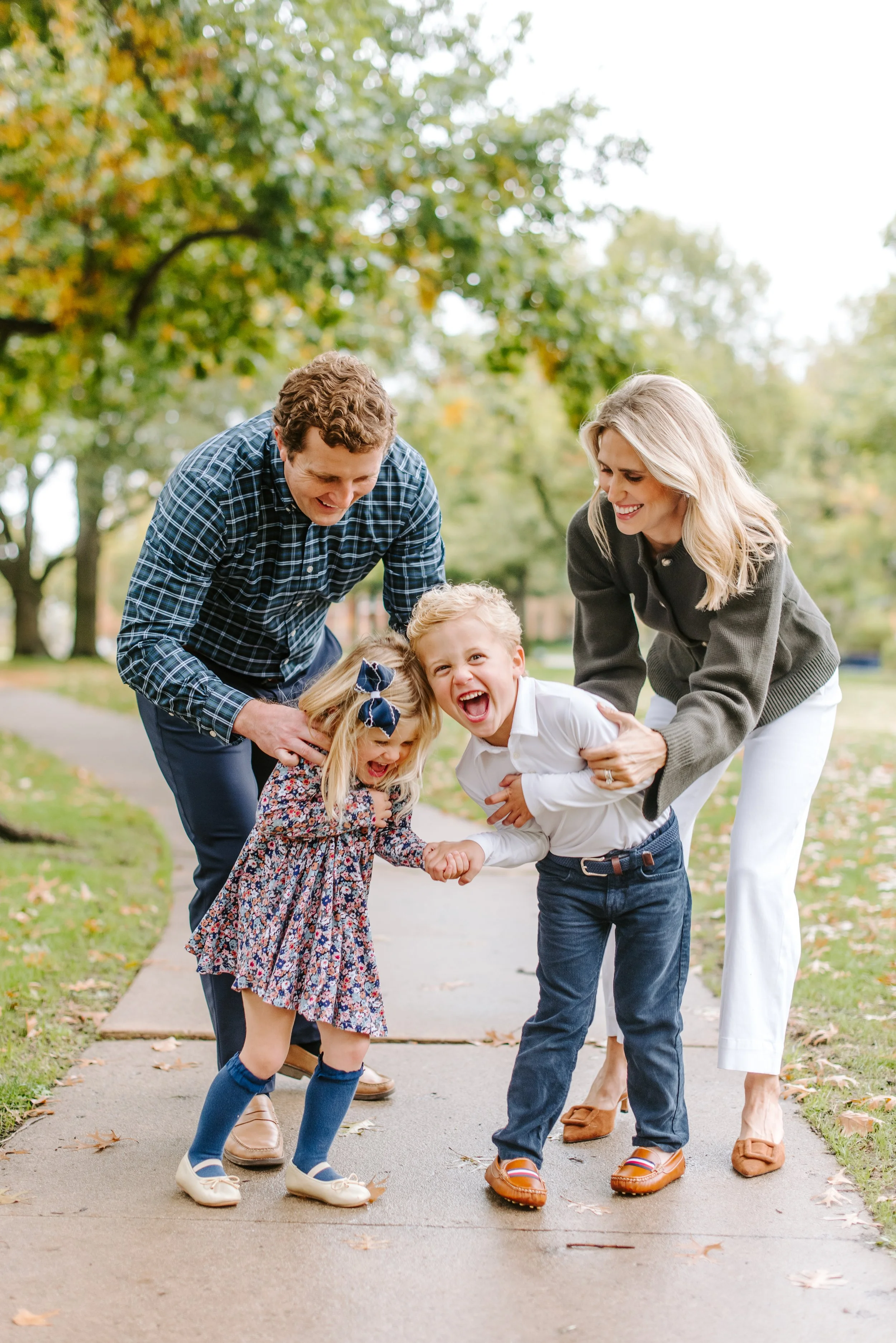 Family of two adults and two children enjoying a playful moment on a park sidewalk, with trees in the background and fallen leaves on the ground.