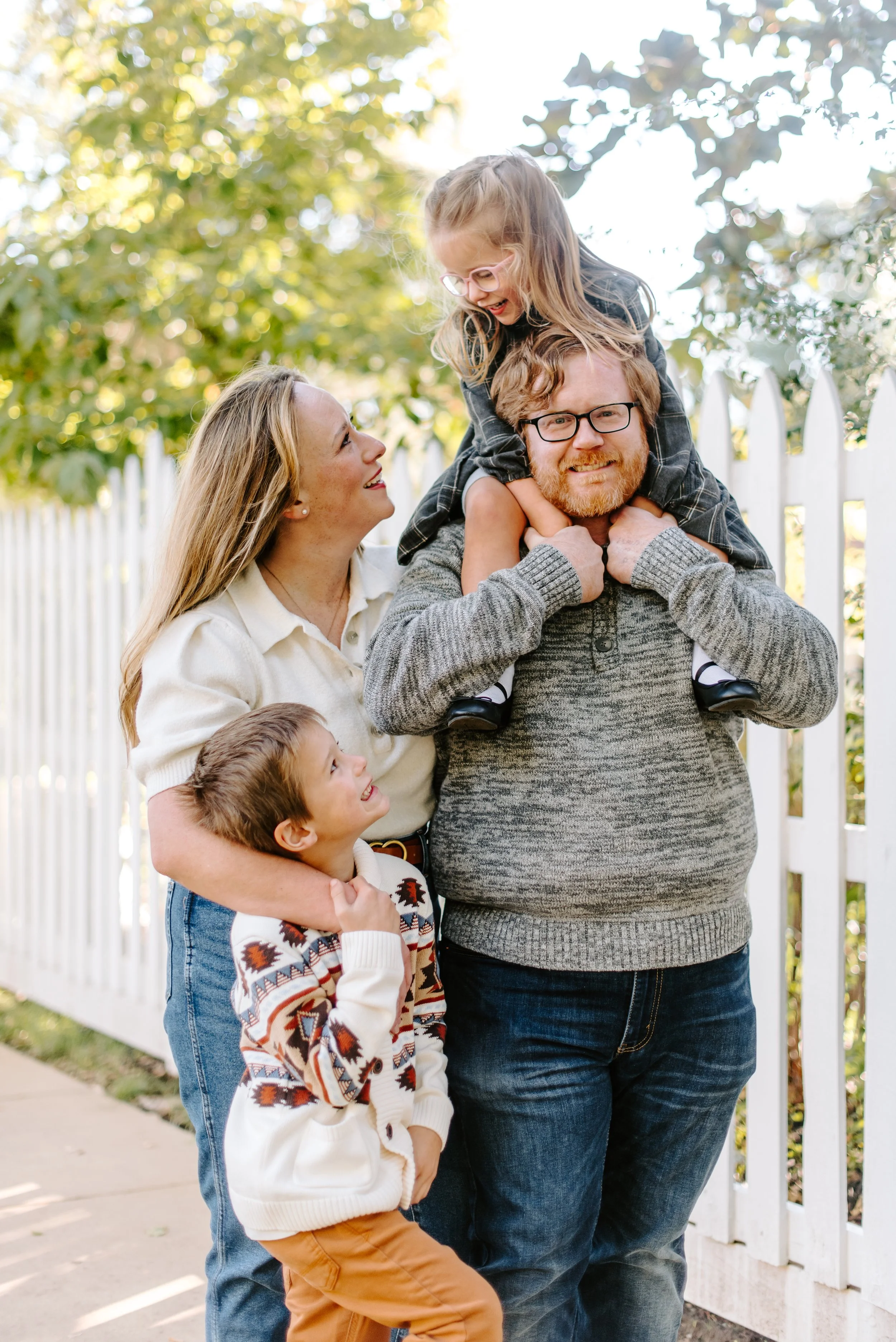 A happy family of five outdoors near a white picket fence. The man is carrying a young girl on his shoulders, and the woman is looking at them. Another young boy is hugging the woman, and a toddler is in the foreground, smiling.