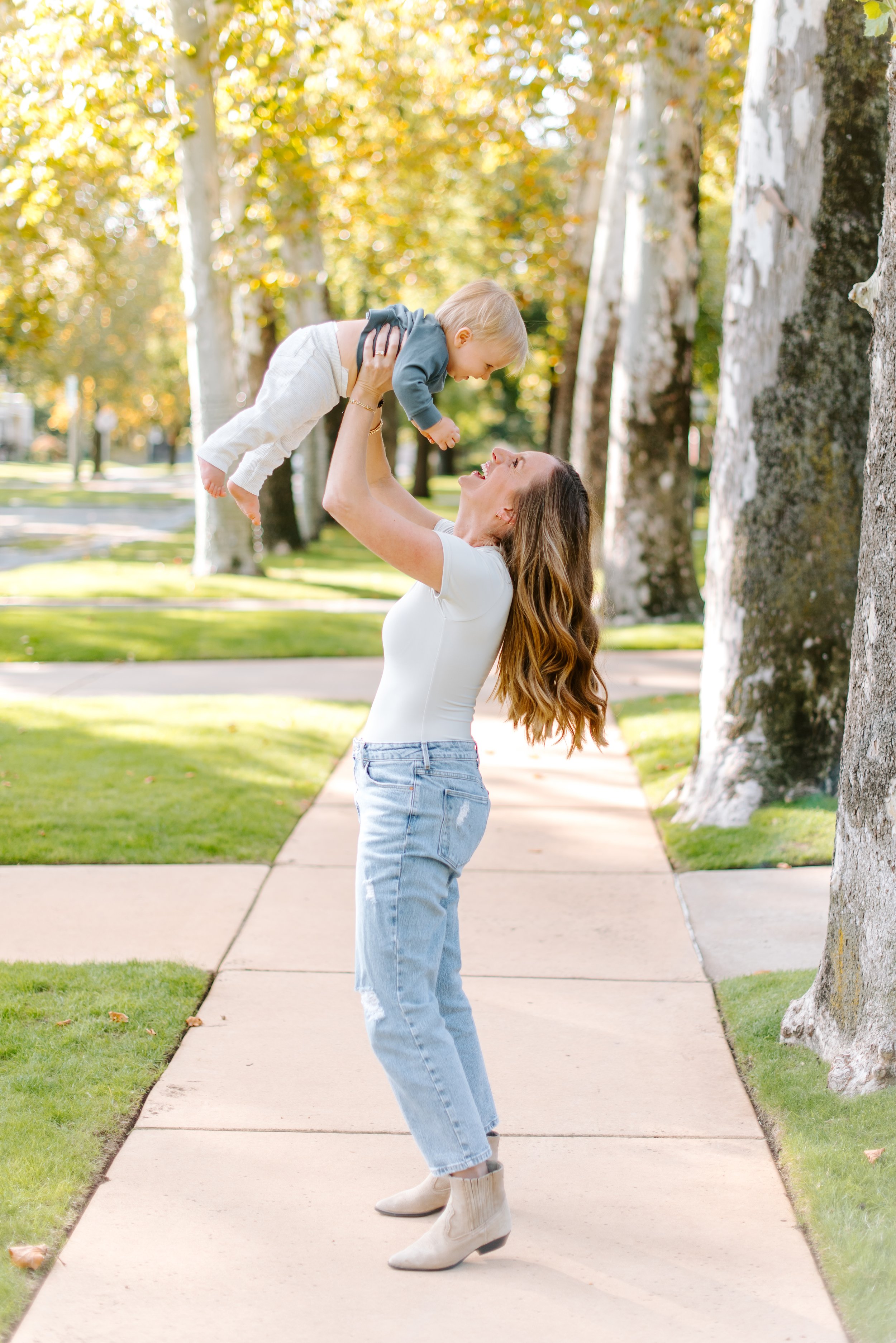 A woman with long, wavy red hair in high-heeled ankle boots, light blue jeans, and a white t-shirt, playing with a toddler boy in a park with trees and a sidewalk.