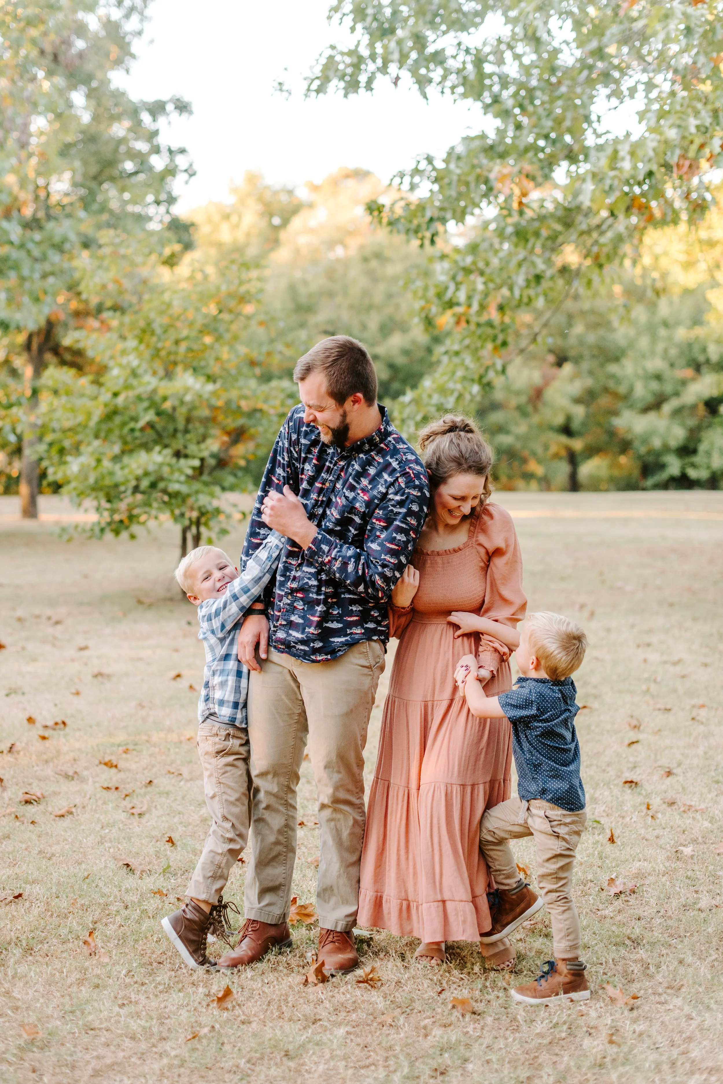A family of five, including two adults and three children, enjoying a day outdoors in a park with green trees and fallen leaves, smiling and interacting playfully with each other.