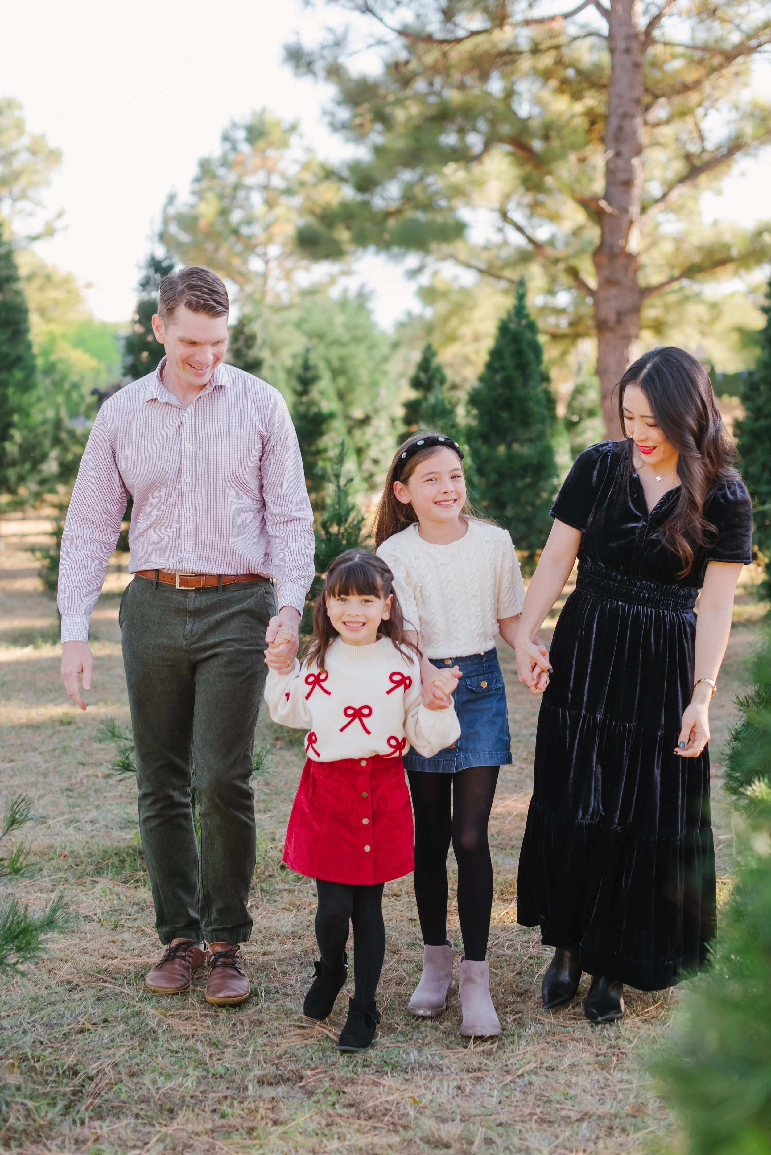 A family of five, including a man, woman, and three young girls, walking outdoors through a grove of pine trees during daytime, all smiling and holding hands.
