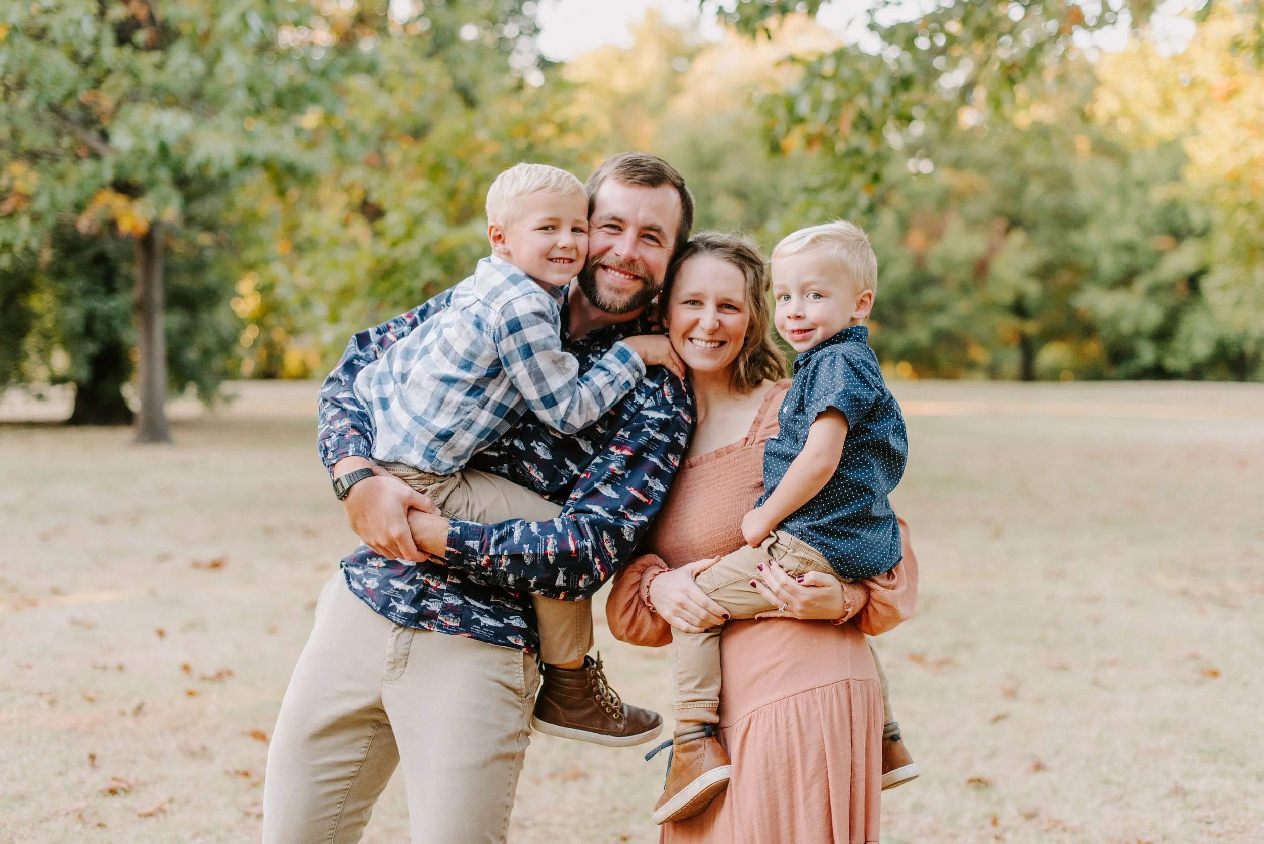 A family of four with two adults and two young children smiling and hugging outdoors in a park with green and yellow foliage in the background.