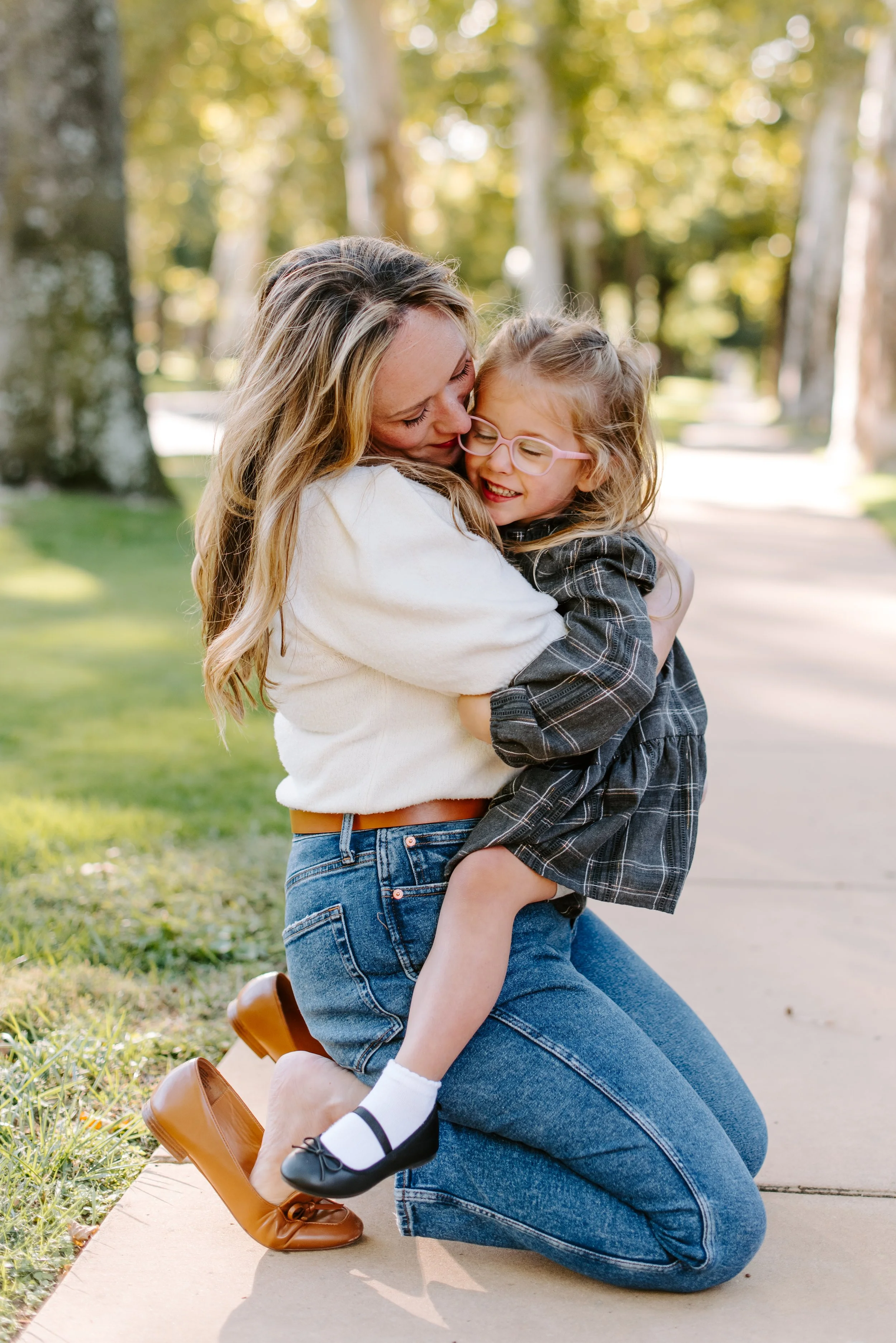 A woman kneeling on the ground embracing a young girl, both smiling and sharing a happy moment outdoors on a sunny day, with trees in the background.