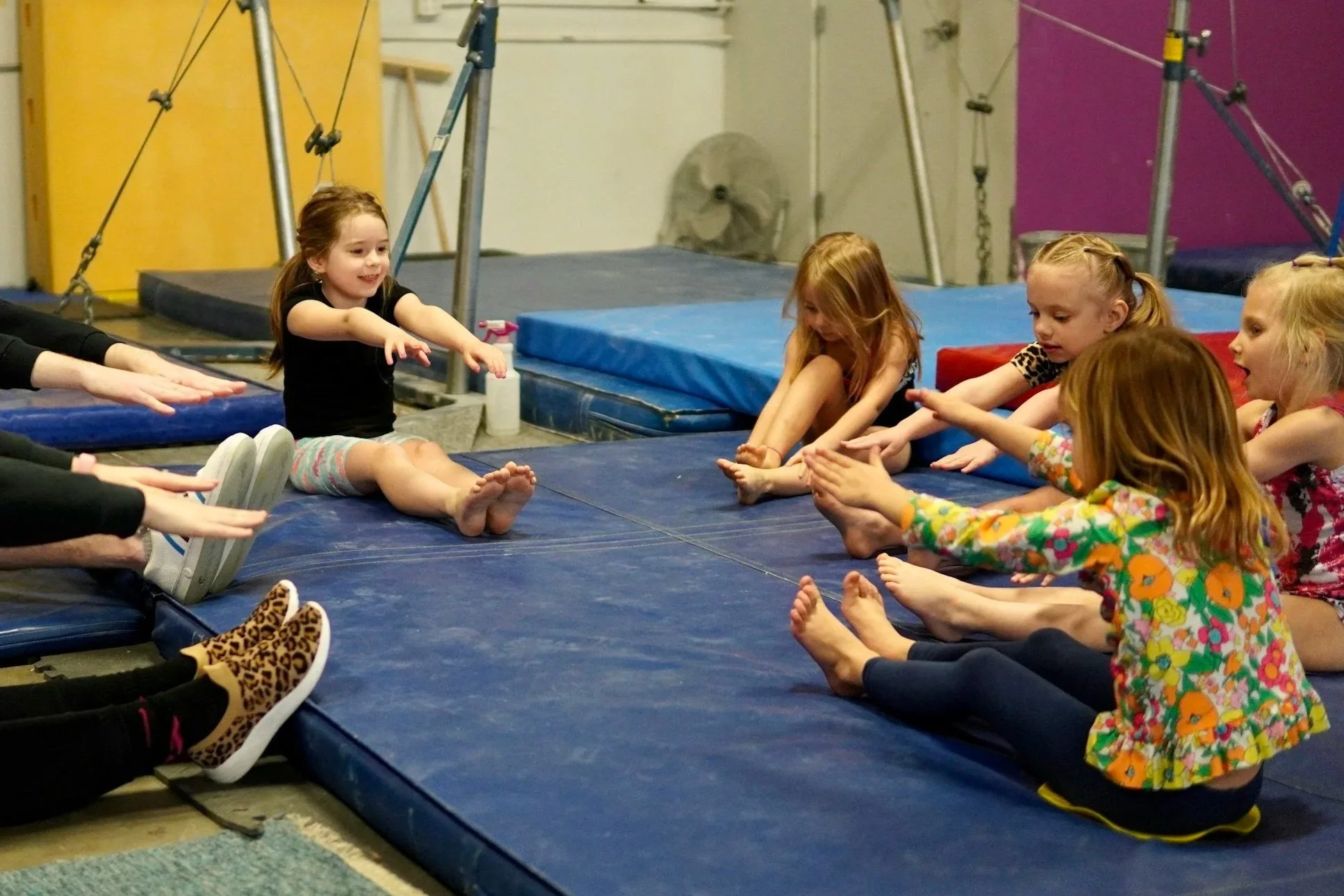 Children sitting on gym mats stretching their legs and reaching forward in a gymnastics class.