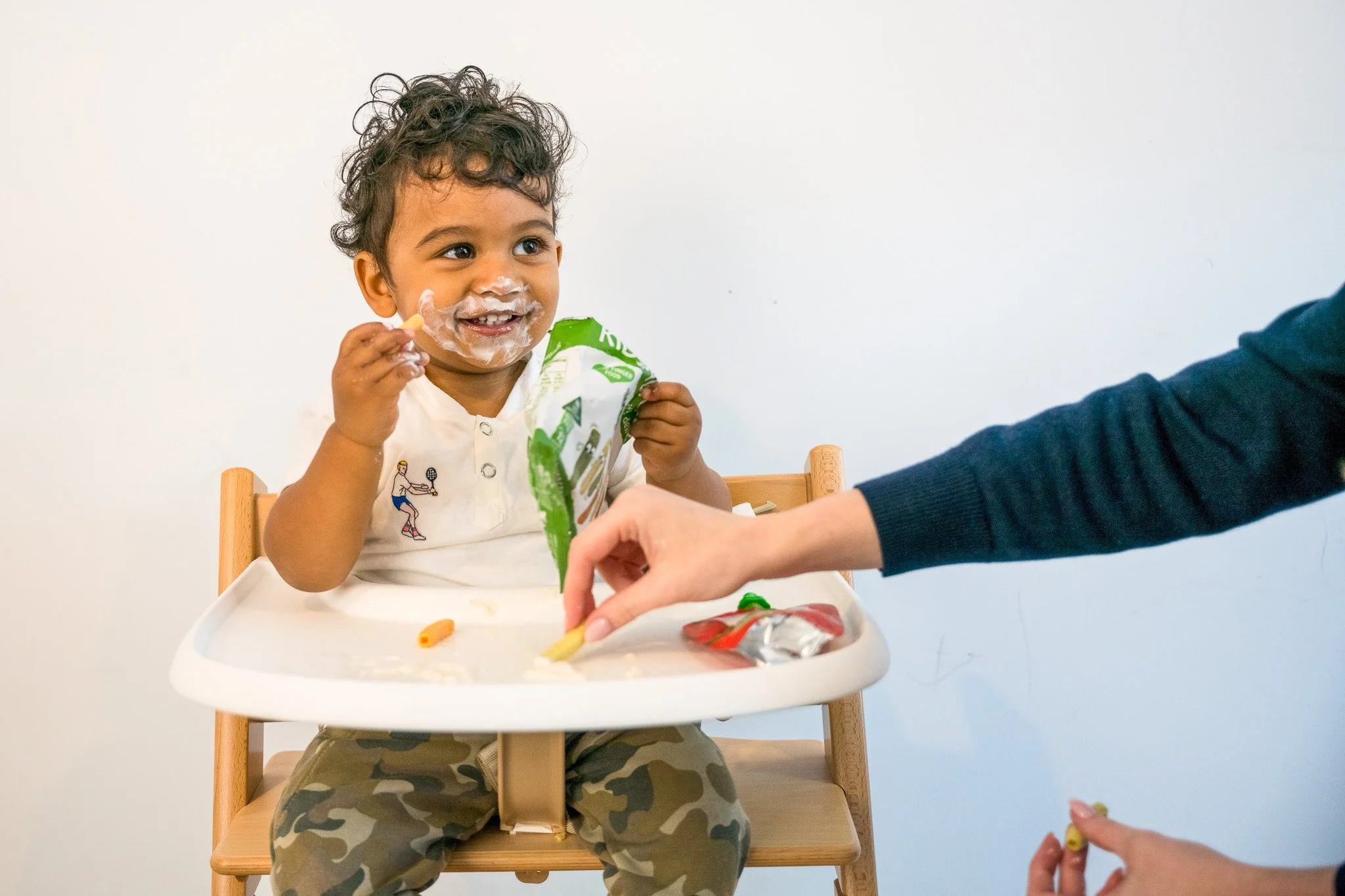 A young boy with curly hair, smiling, sitting in a high chair with messy food on his face, eating snacks handed to him by an adult whose arm is visible. Showing Therapy for picky eating / mealtime difficulties