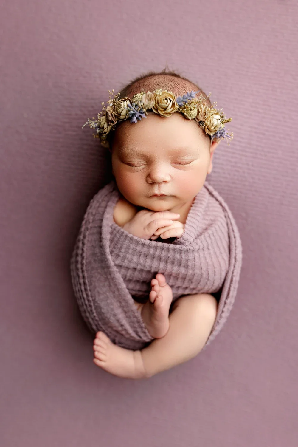 Sleeping newborn baby girl wrapped in soft purple fabric wearing delicate floral headband posed on textured backdrop