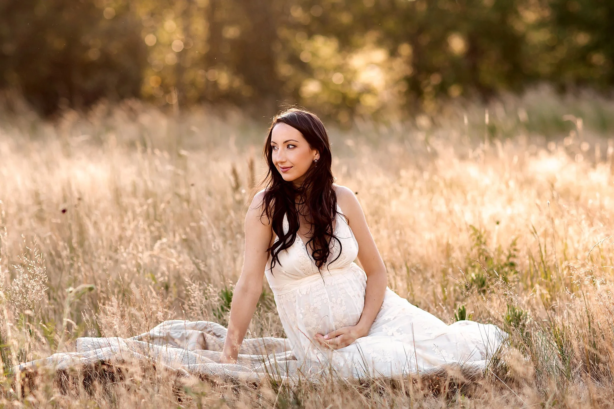 Pregnant mother seated in golden field wearing white dress during sunset maternity session in Salem Oregon