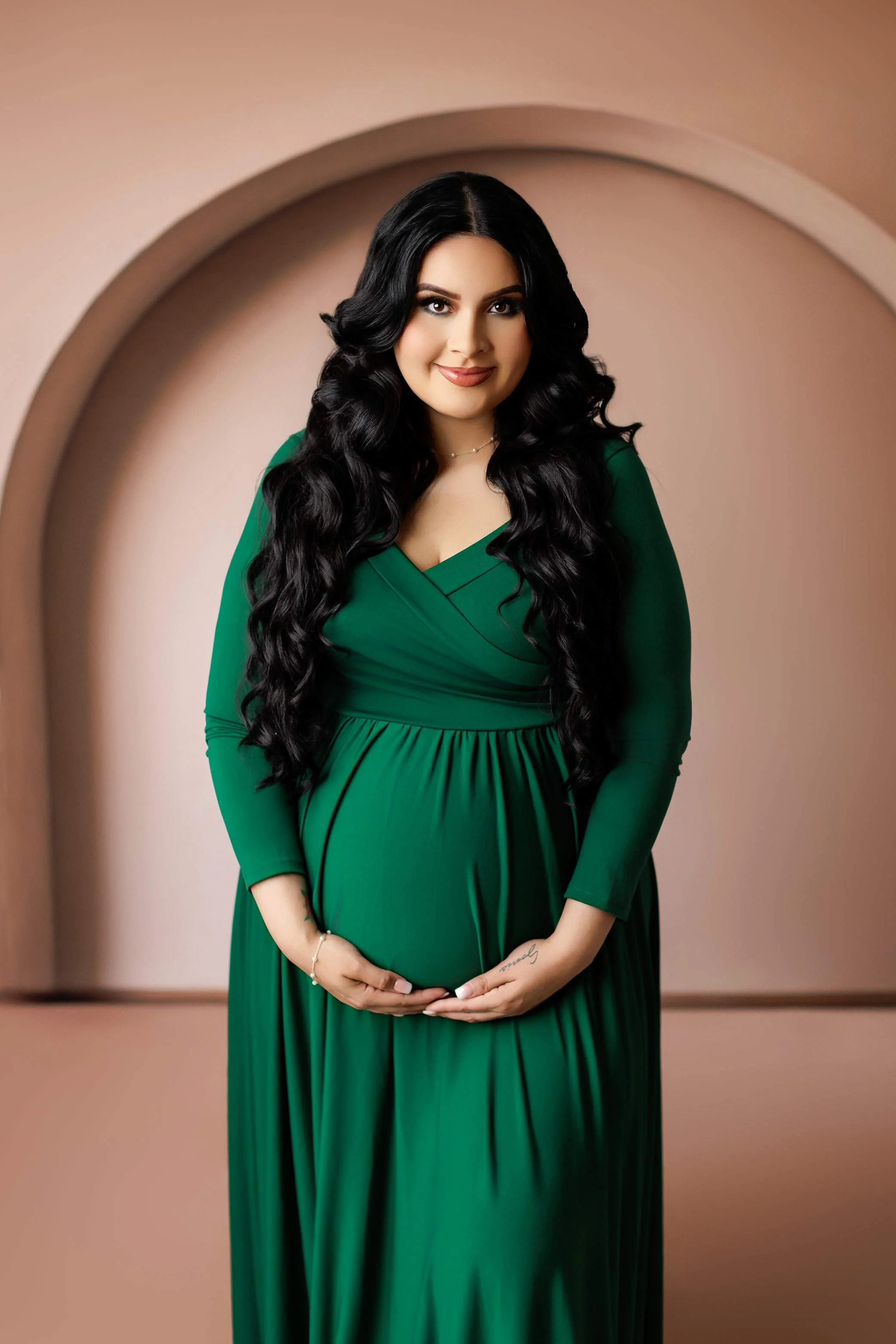 Pregnant woman in green dress posing in studio, elegant maternity portrait with soft neutral arch backdrop