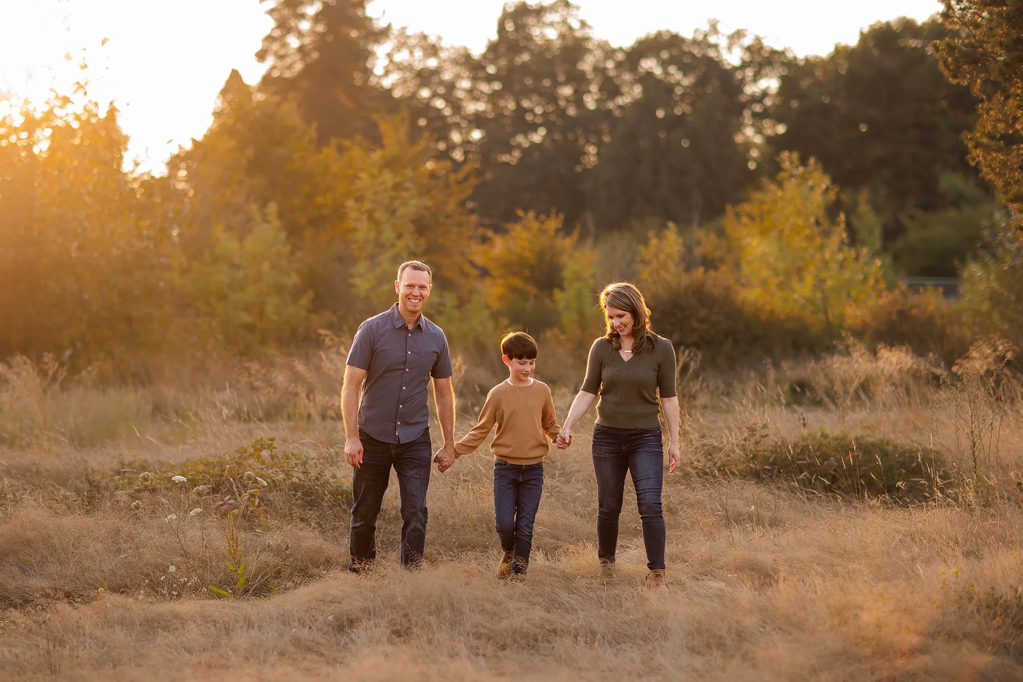 Family of three walking through golden field with child in the middle holding parents’ hands.