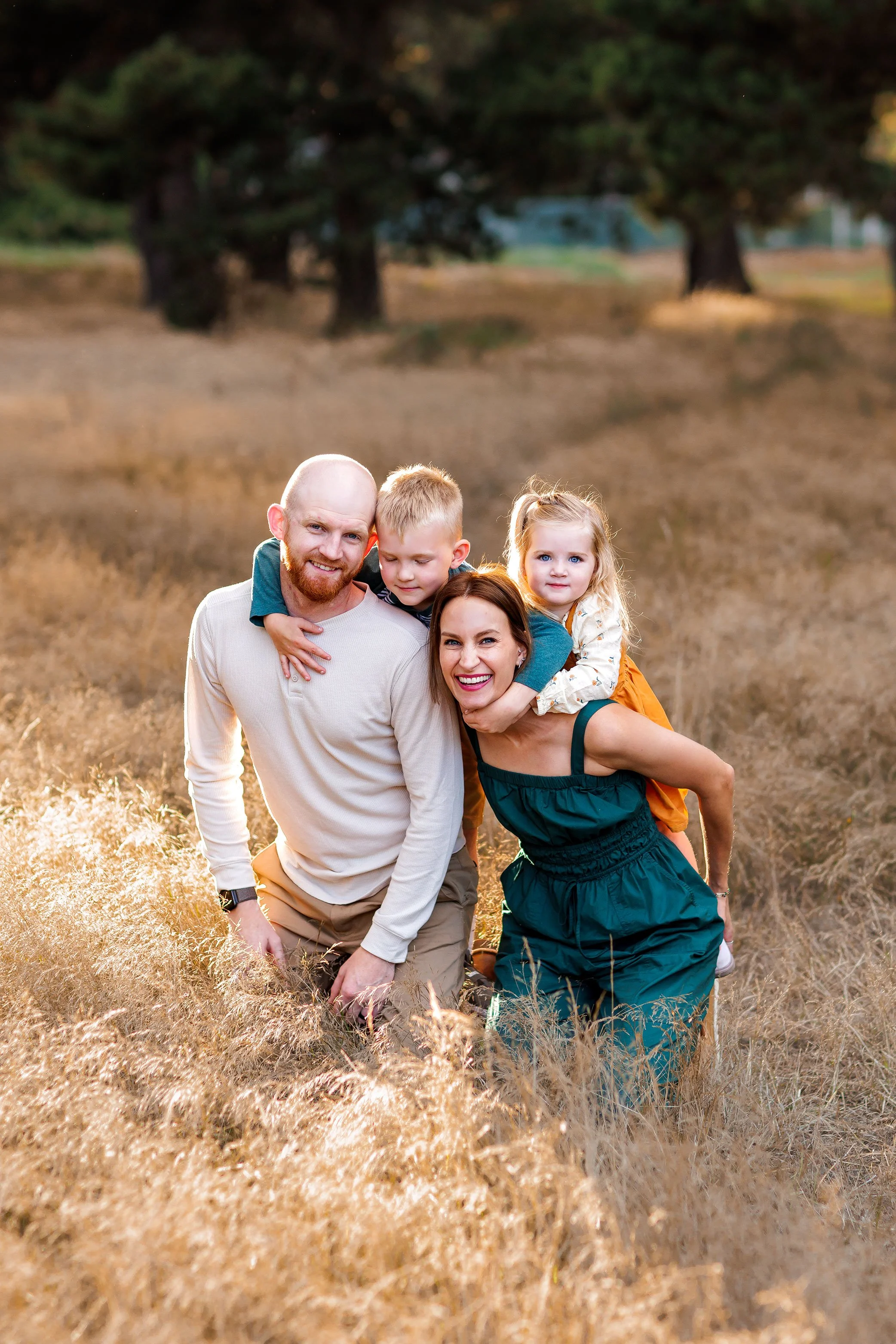 Family of four kneeling in golden field while children climb onto parents’ backs during playful outdoor session.