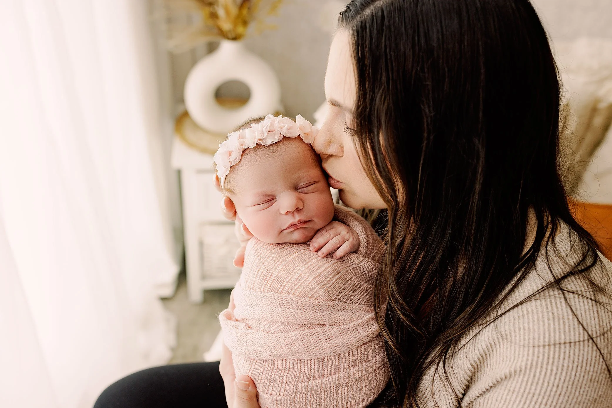 Mother kissing her sleeping newborn baby girl wrapped in soft pink fabric near a window.