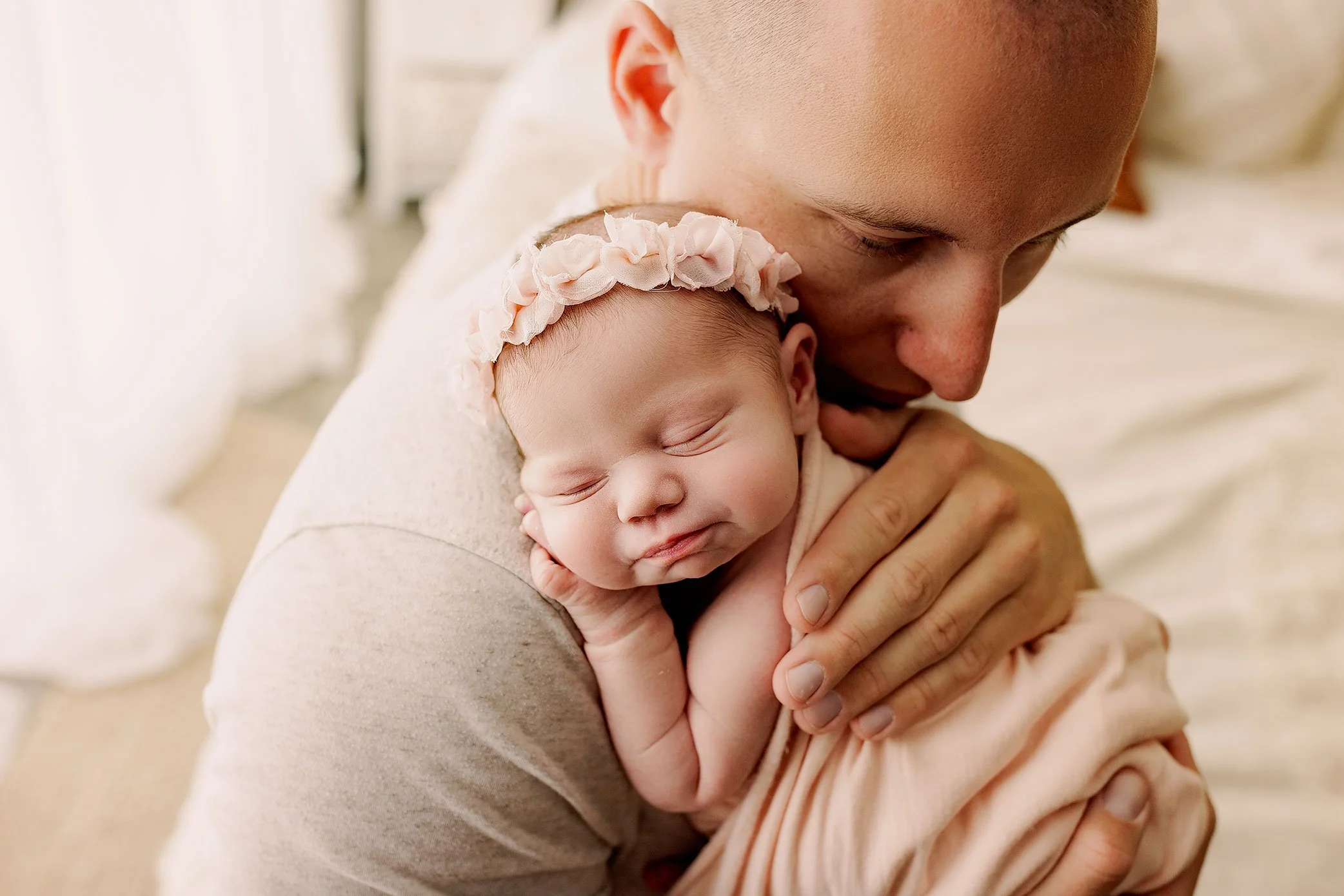 Sleeping newborn baby girl with a soft floral headband resting peacefully on her father’s shoulder.