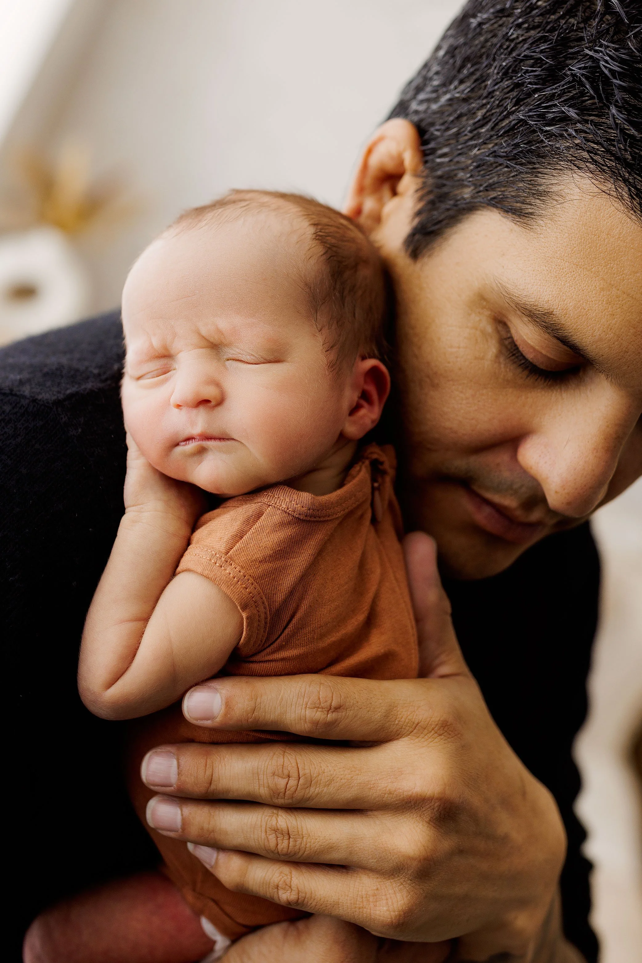 Sleeping newborn baby resting against father’s shoulder in a warm, vertical, close-up portrait.