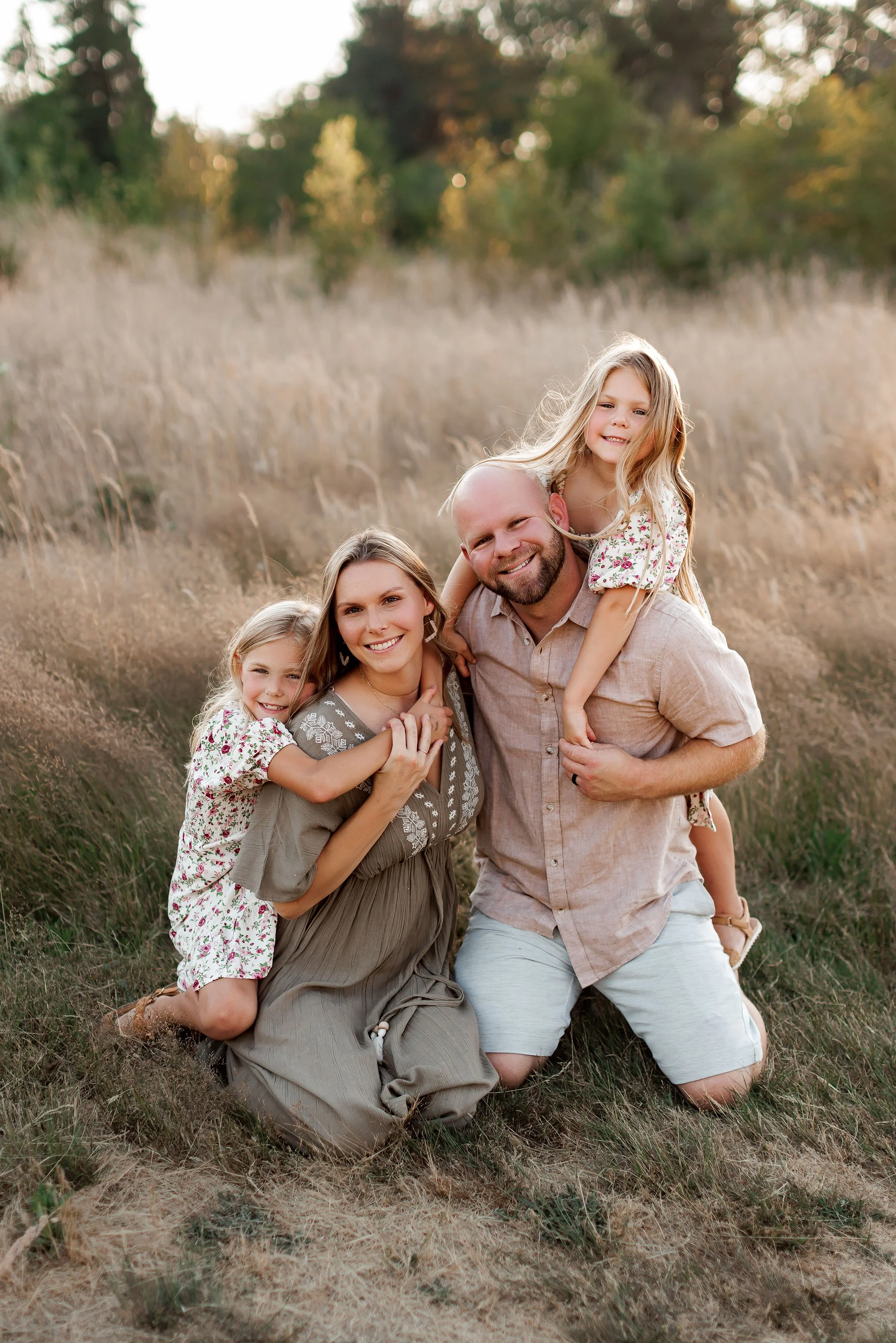 Family of four outdoors at golden hour with parents kneeling at different heights and daughters hugging them.