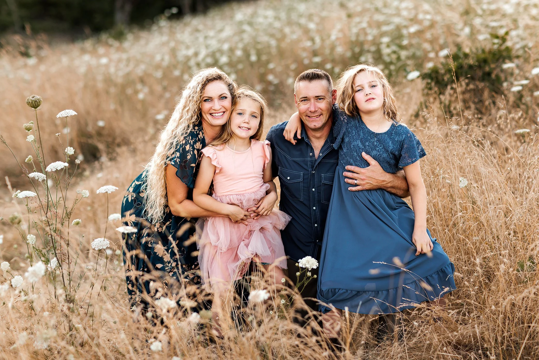 Family of four kneeling in tall grass at golden hour with parents positioned to align faces in a row.