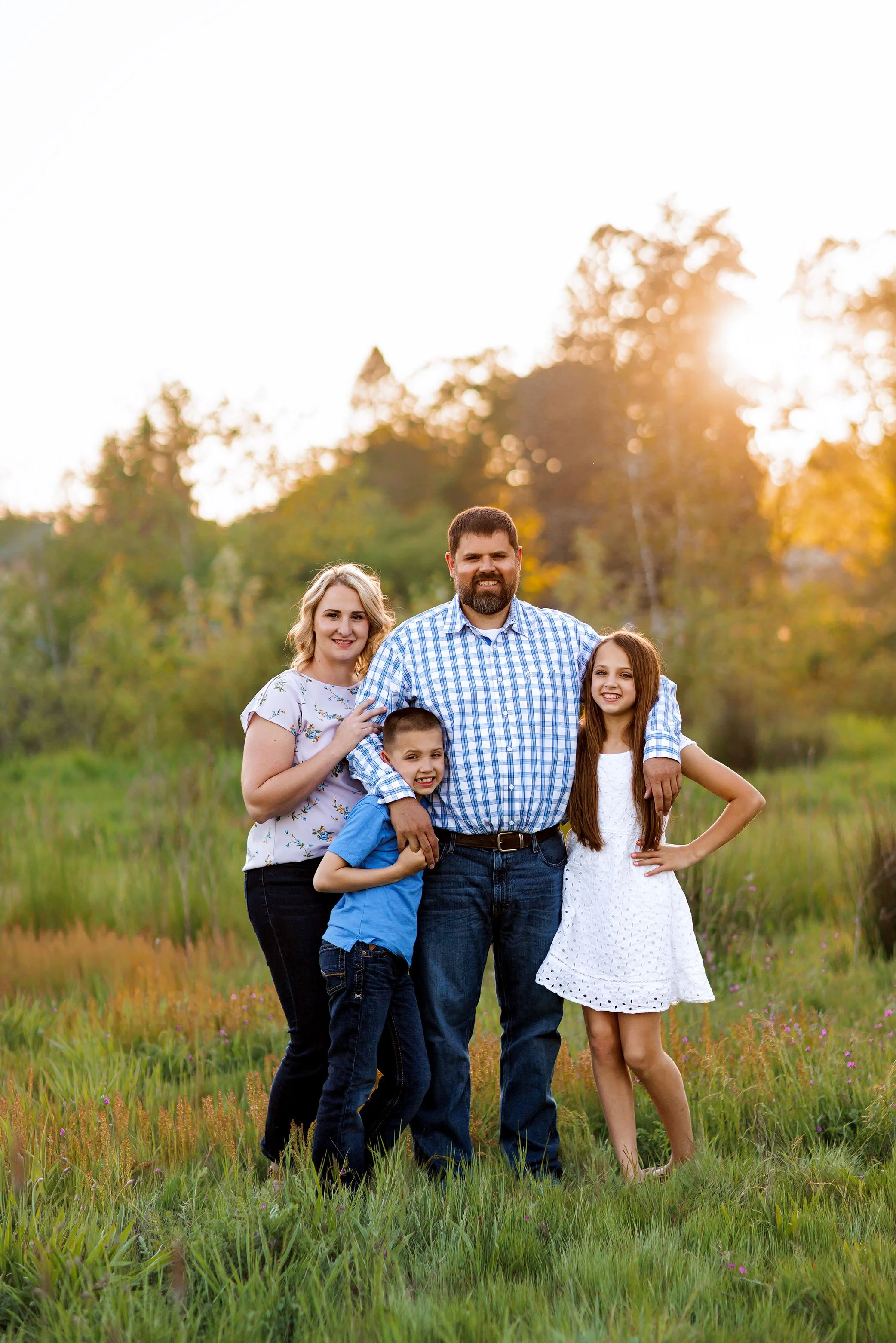 Family of four standing in field at sunset with dad’s arms around children and mom leaning into him.