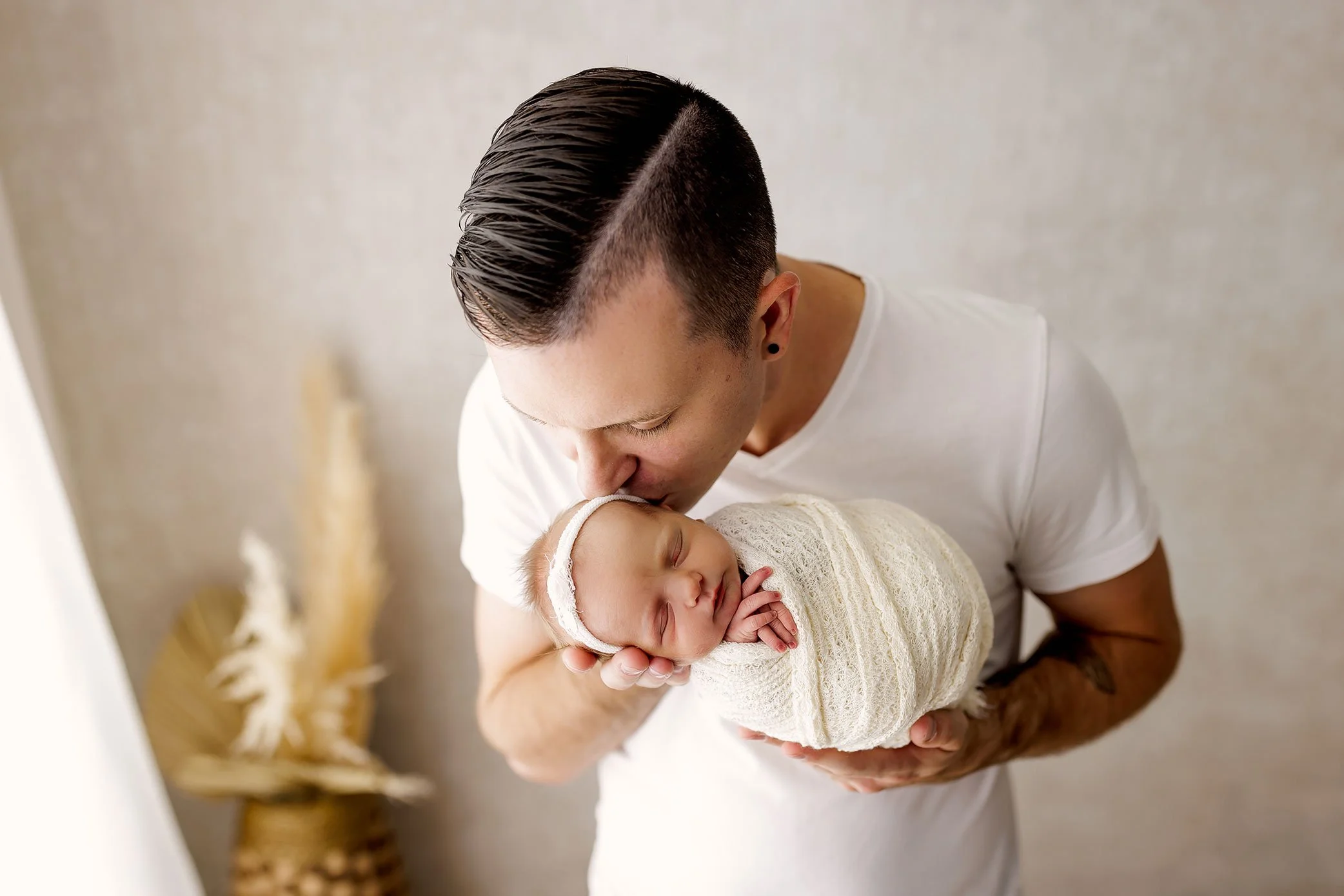Father gently kissing his swaddled newborn baby’s forehead in a soft, neutral studio setting.
