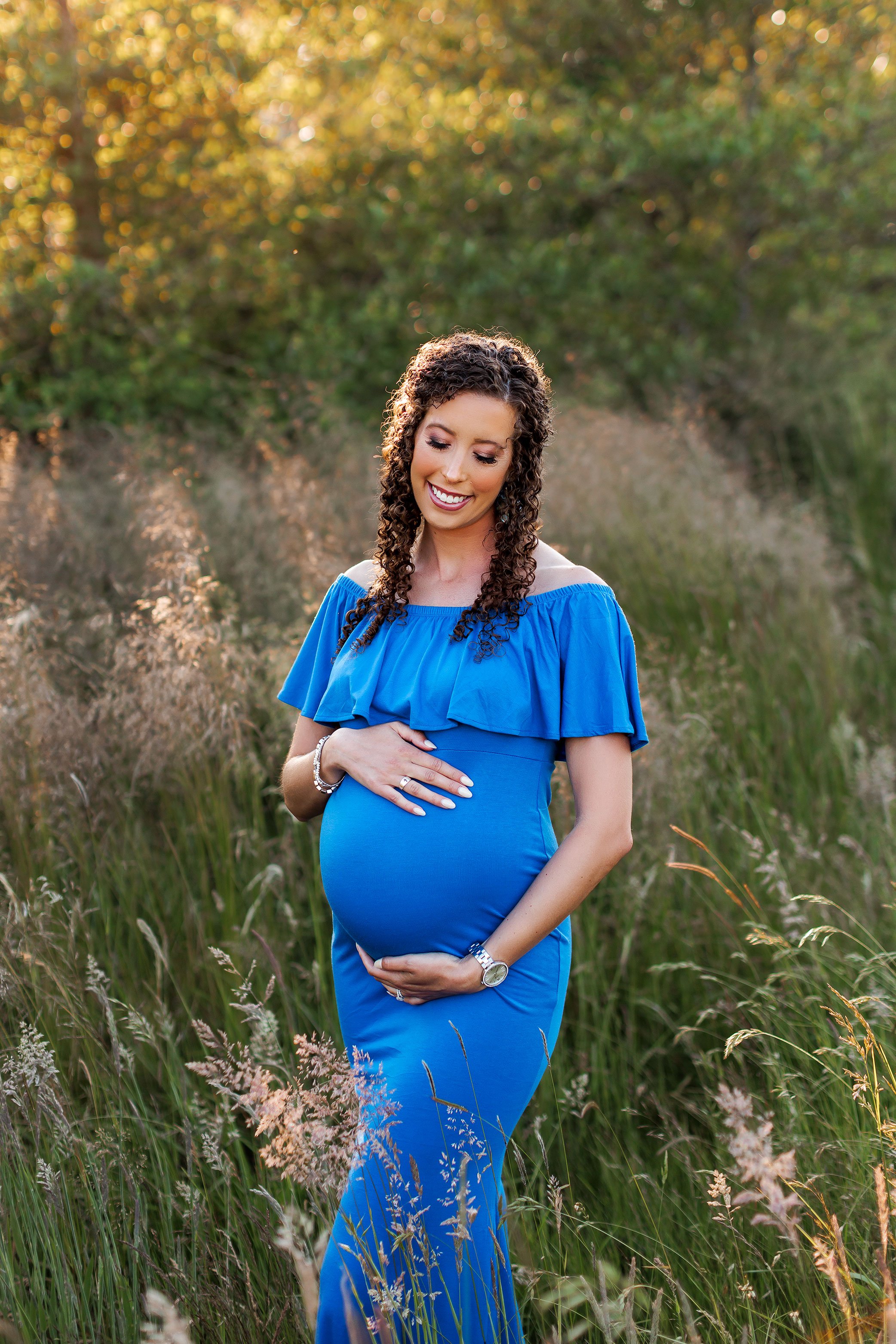 Pregnant mother in fitted blue gown standing in tall grass during outdoor maternity session.