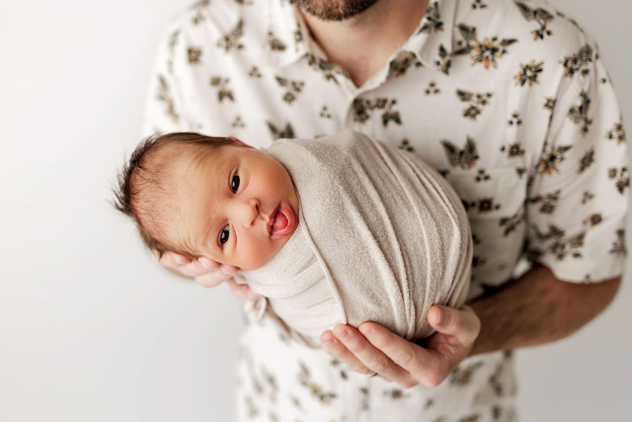 Newborn baby boy wrapped in a soft beige swaddle, sticking his tongue out while being held.