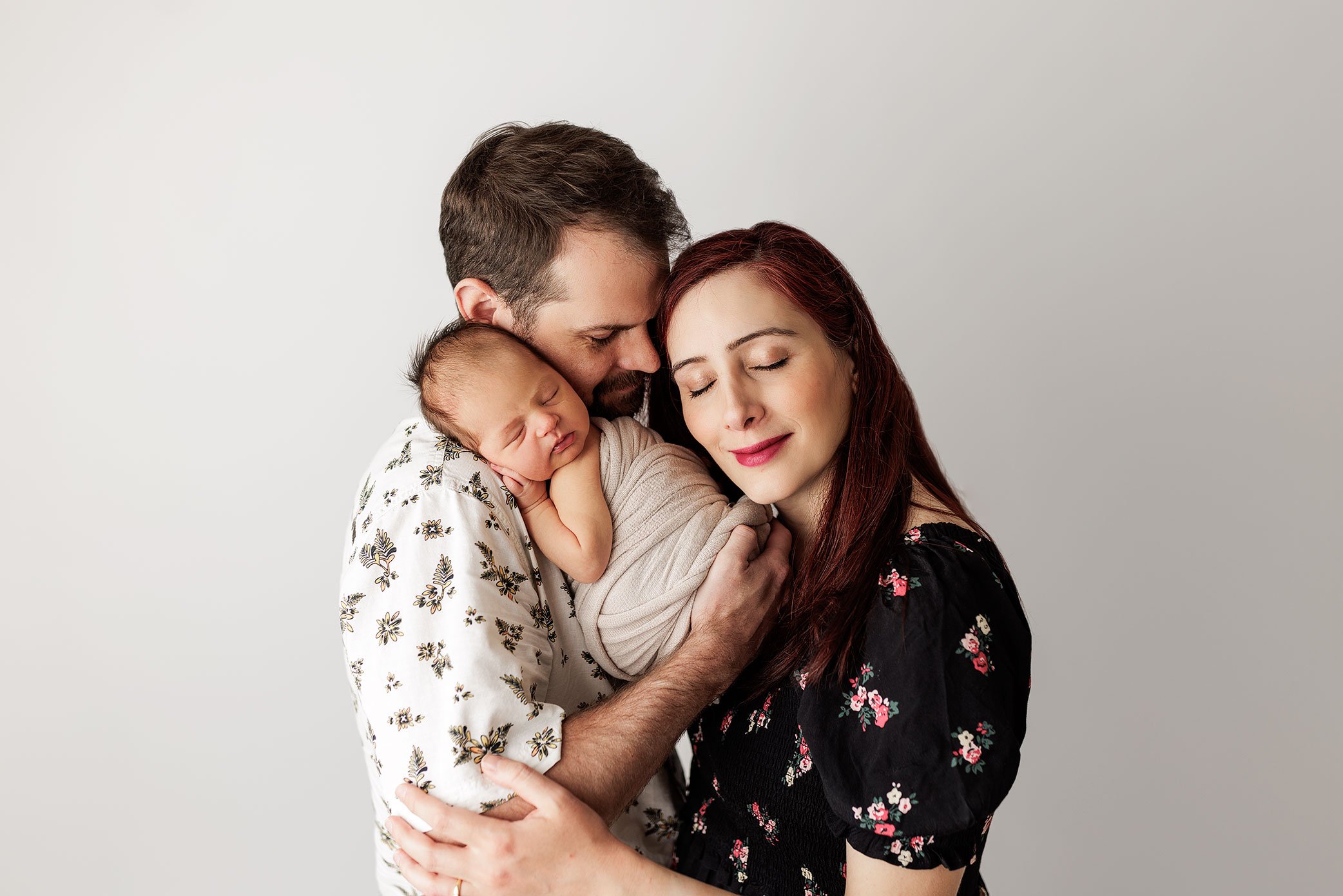Mother and father holding their sleeping newborn baby in a soft, neutral studio portrait.