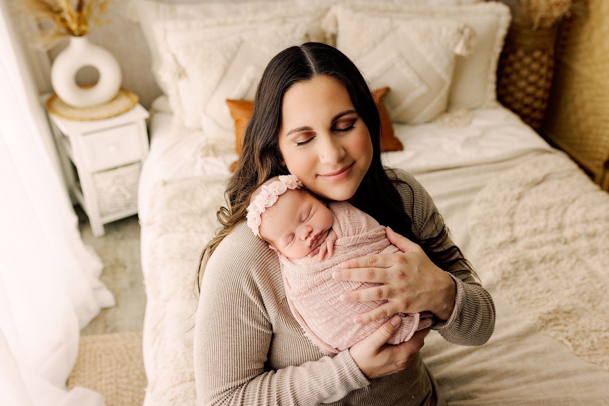 Mother holding her sleeping newborn baby girl wrapped in soft pink fabric in cozy bedroom setting.